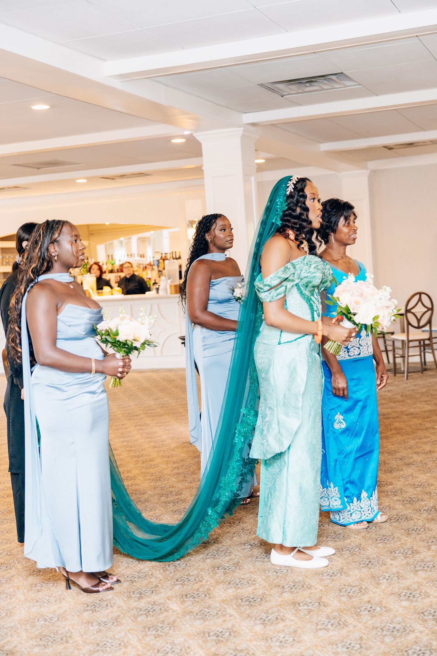 Bridesmaids in light blue gowns stand with bride in teal traditional African wedding attire.