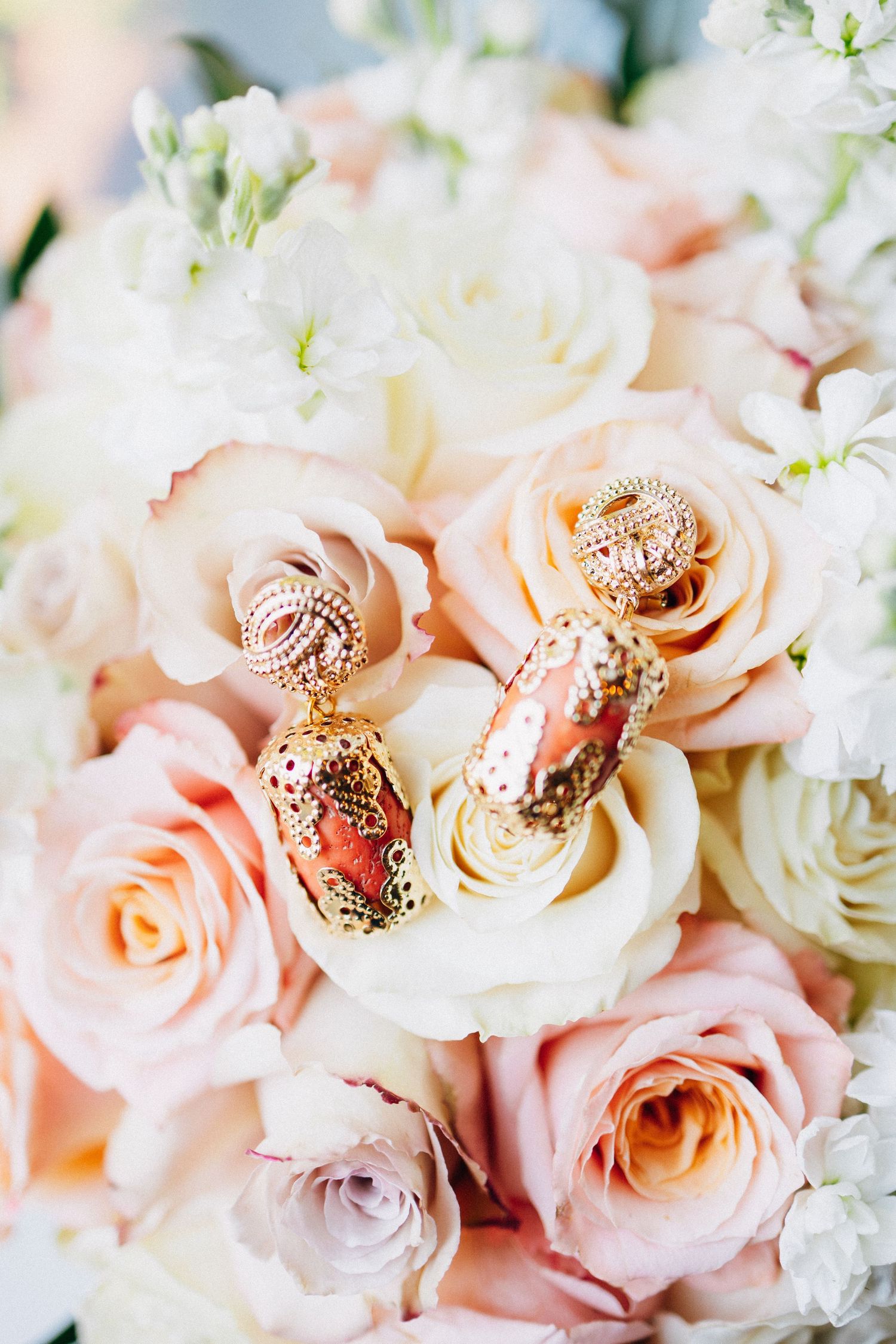 Gold and coral drop earrings resting on a bridal bouquet of blush pink and white roses.