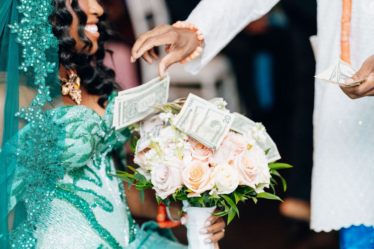 Close-up of bride in teal sequin dress receiving money on bouquet of pink roses at traditional African wedding.