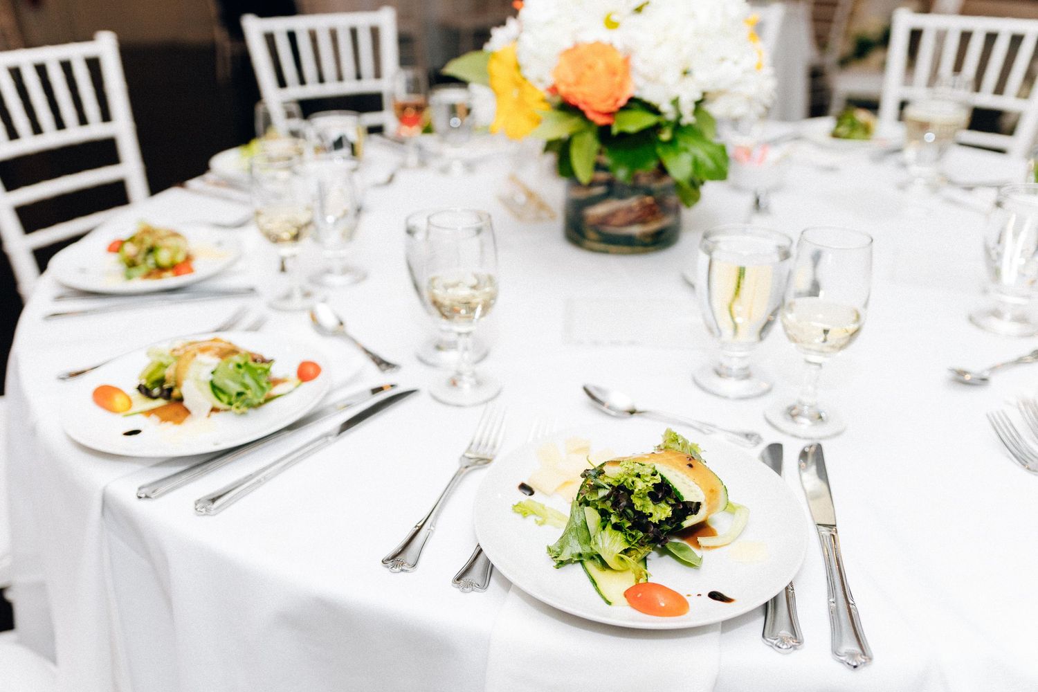 Elegant wedding reception table with white linens, plated salad appetizers, wine glasses, and a floral centerpiece.