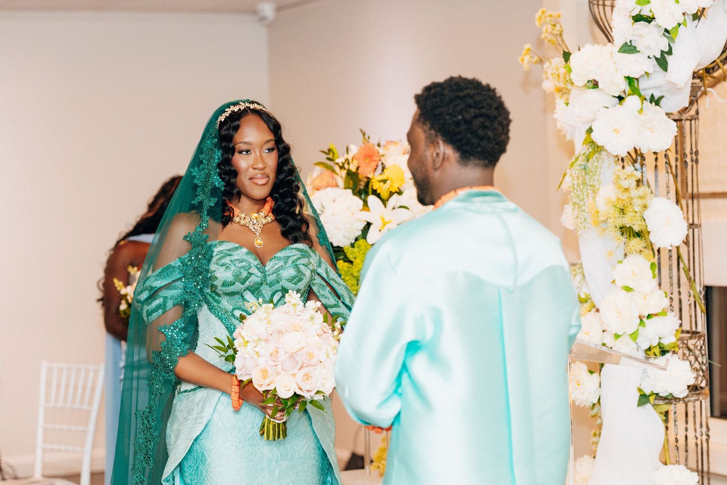 Bride in green gown holds bouquet as groom in mint suit holds her hand at floral wedding ceremony.