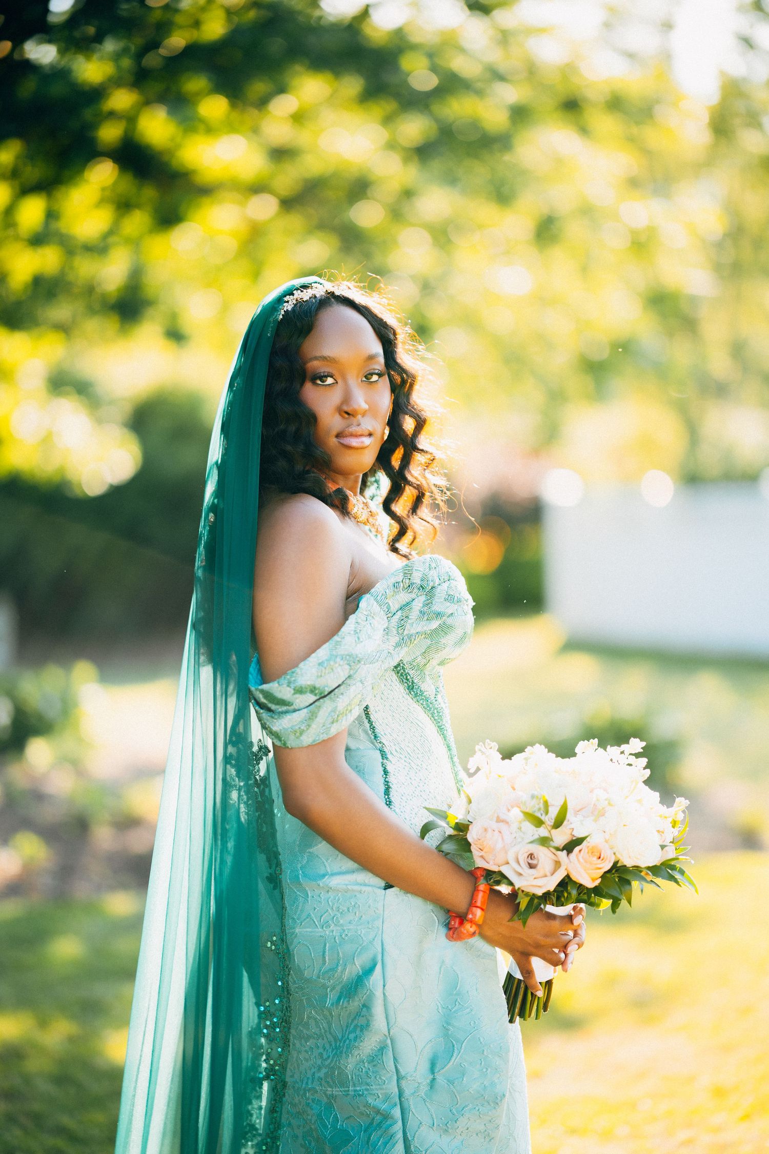 African bride in teal gown and veil holds white bouquet outdoors.