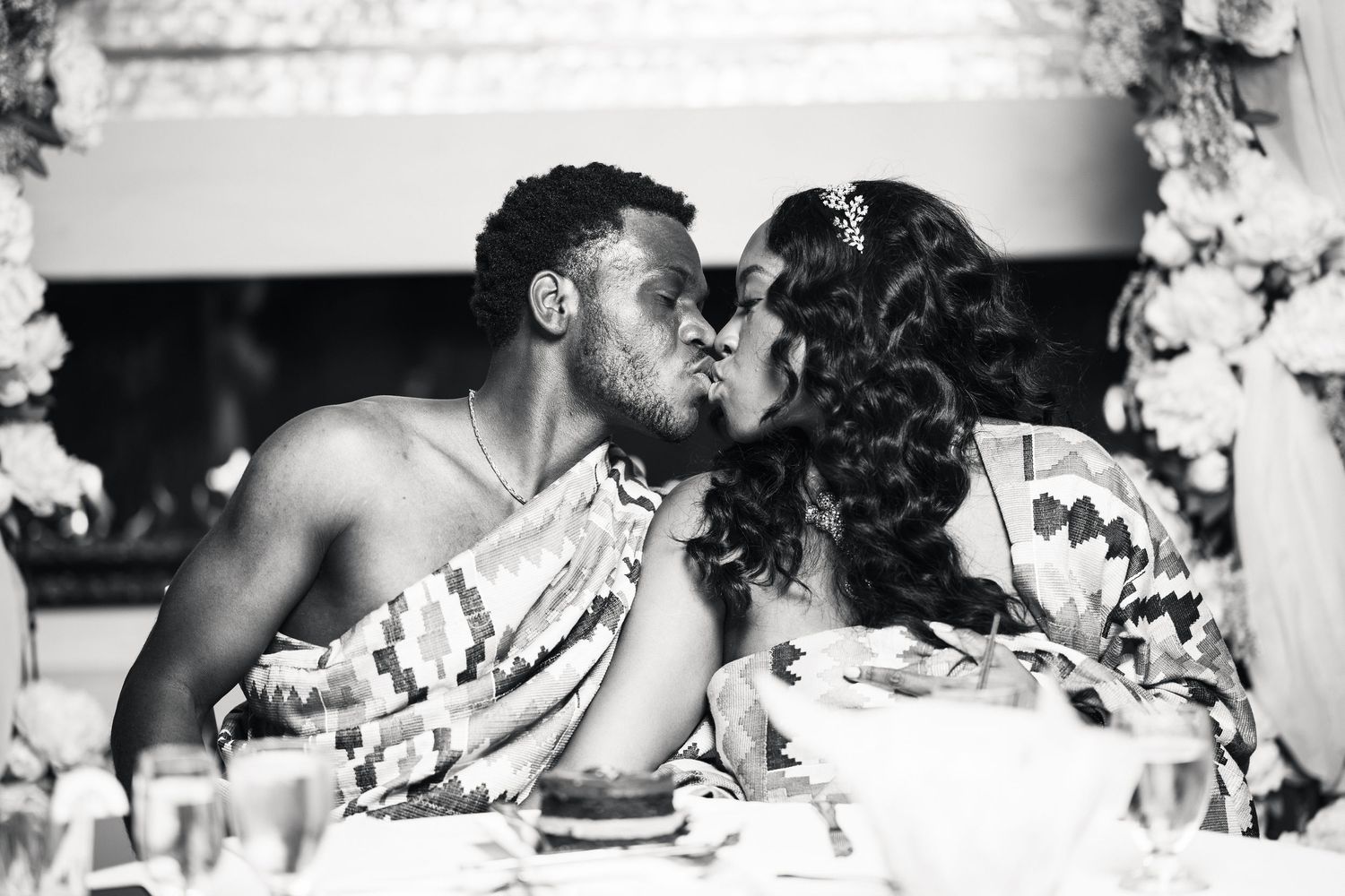A black and white photo of a couple sharing a romantic kiss at their traditional African Kente wedding.