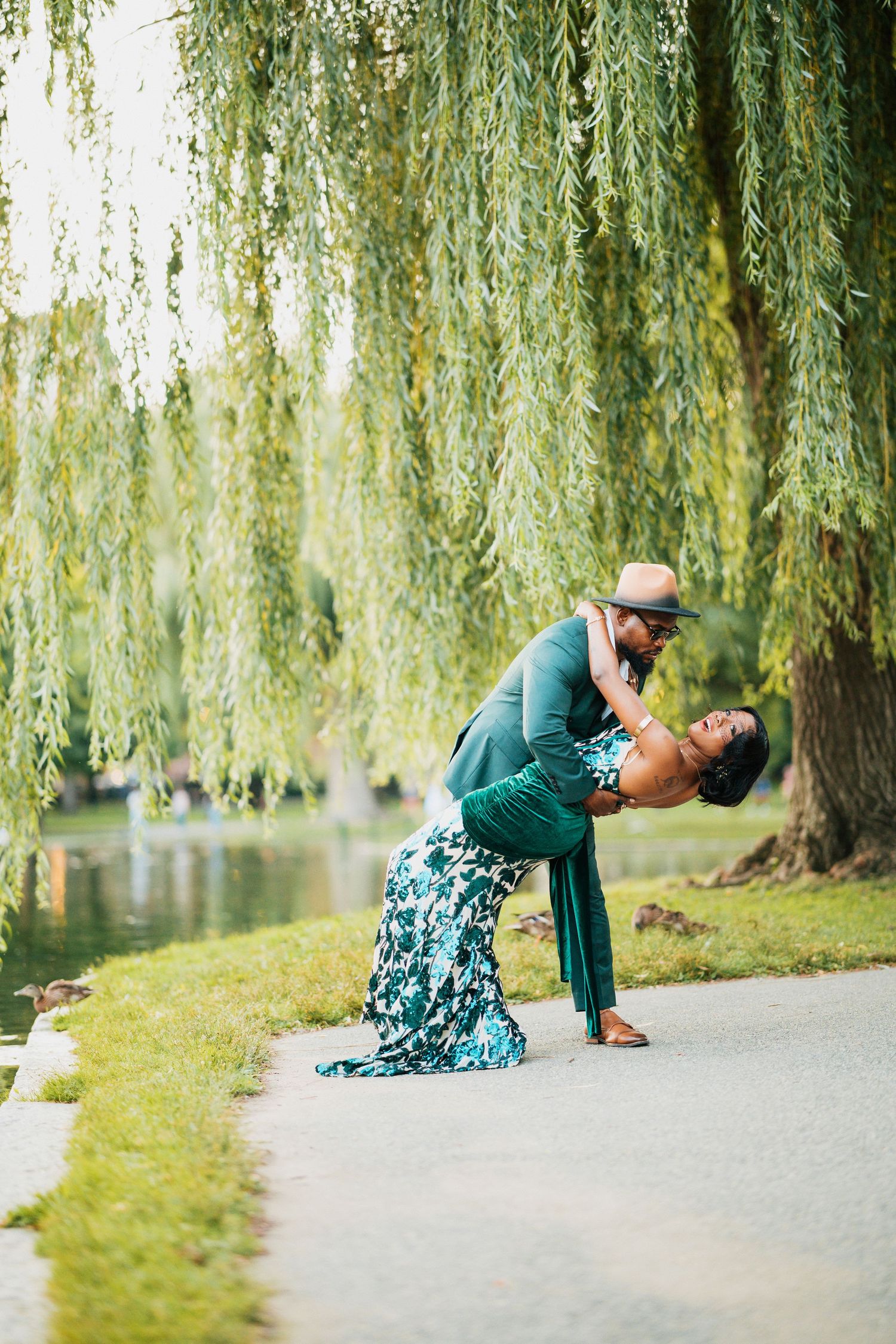 Couple sharing a romantic dip kiss under a weeping willow tree by a pond, wearing coordinated green outfits.