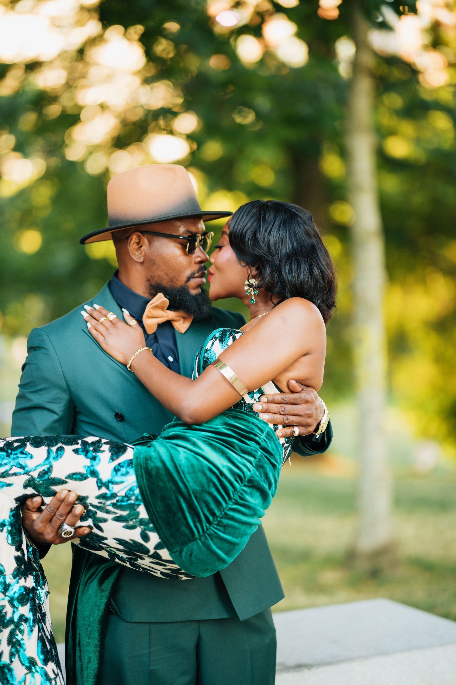 Stylish couple kissing outdoors, man in green suit and tan hat, woman in green velvet and floral dress.