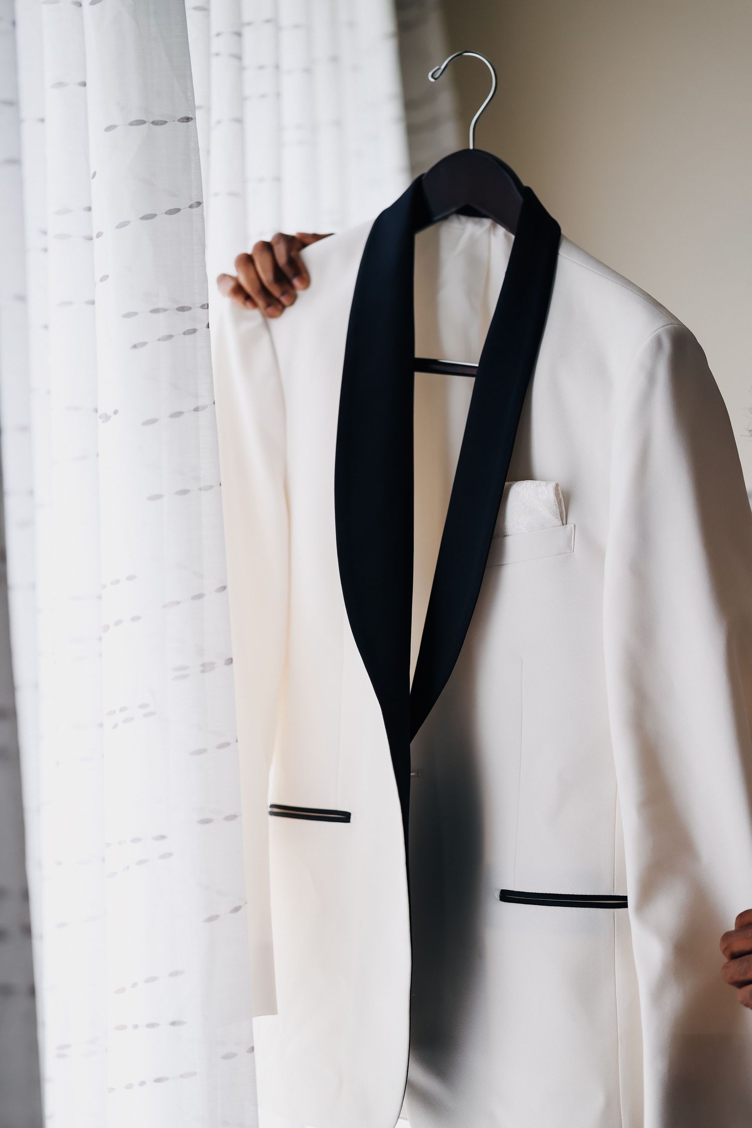 Elegant white tuxedo jacket with black lapels hanging on wooden hanger near curtained window.