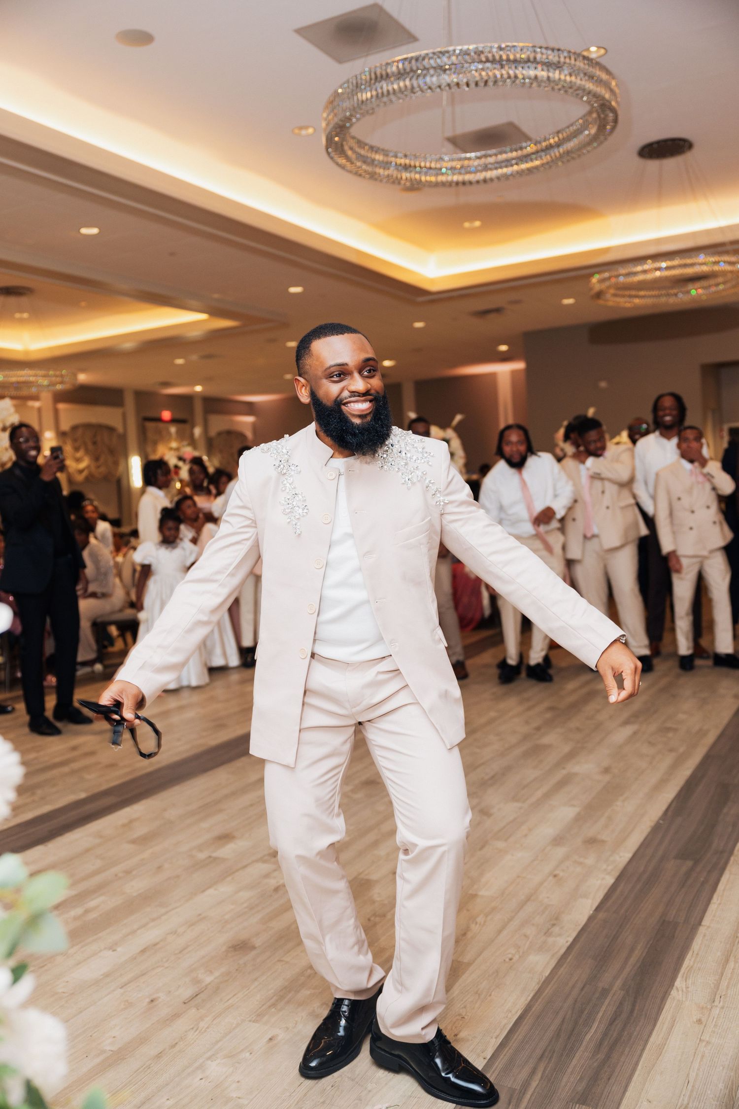 A joyful groom in a cream suit dances at his wedding reception in an elegant ballroom with guests seated behind him.