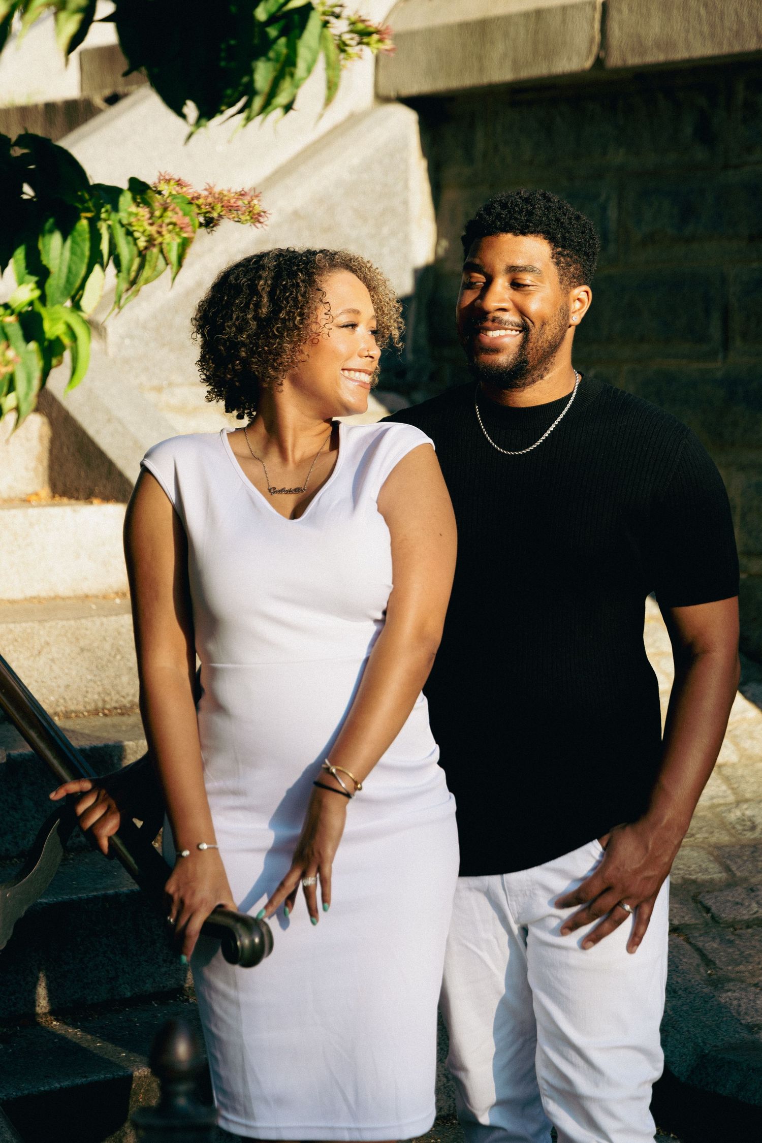 A smiling Black couple in white and black outfits stand together on stone steps surrounded by greenery.