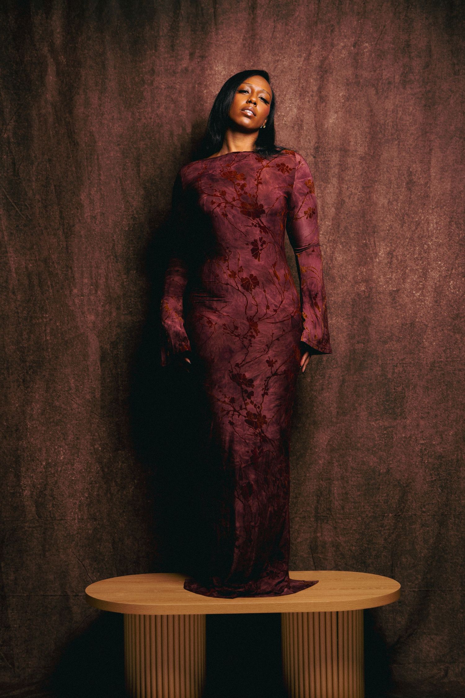 A woman in a fitted burgundy lace gown stands on a wooden platform against a dark mauve backdrop.