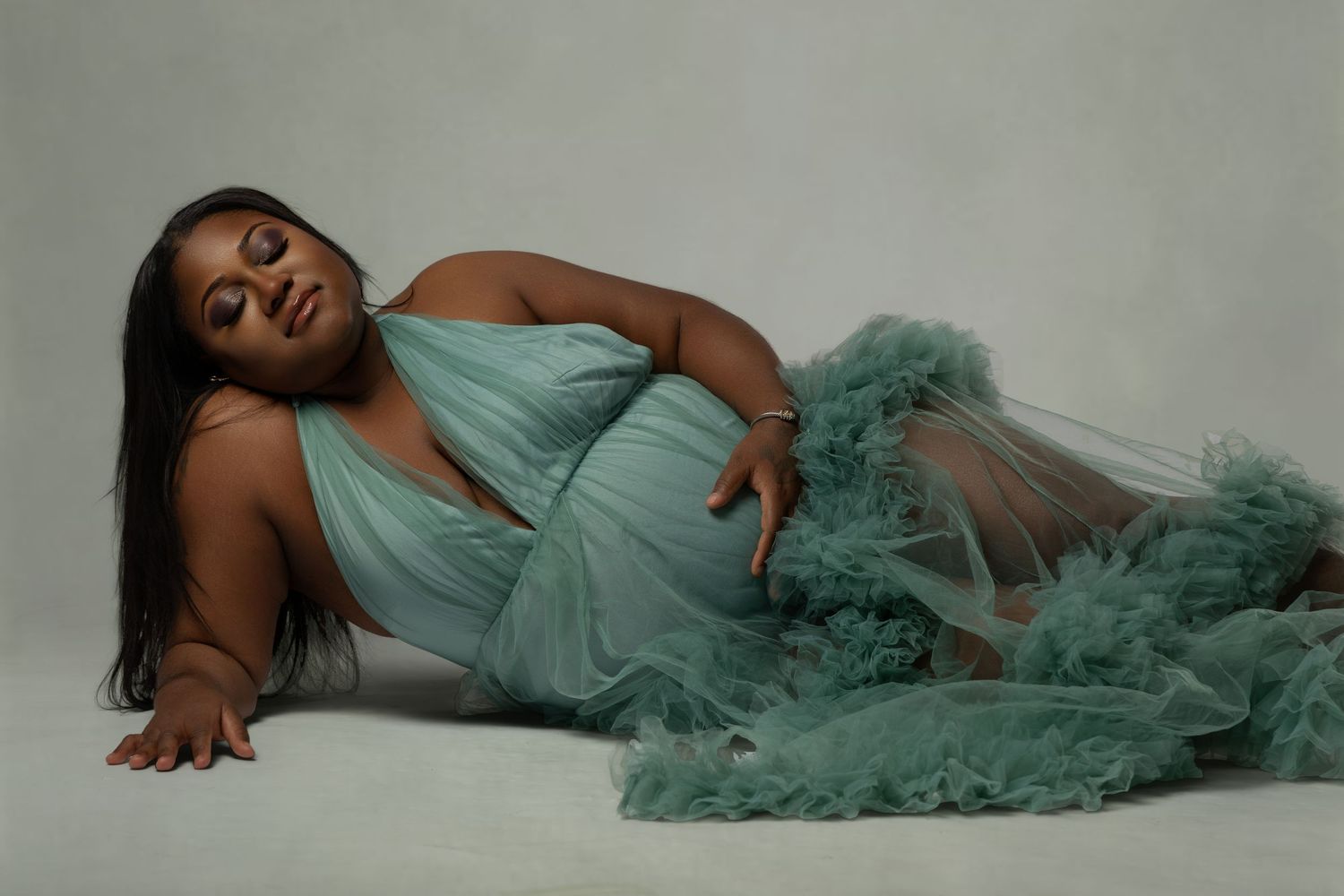 Pregnant woman in mint green tulle gown poses elegantly on floor in professional maternity photoshoot.