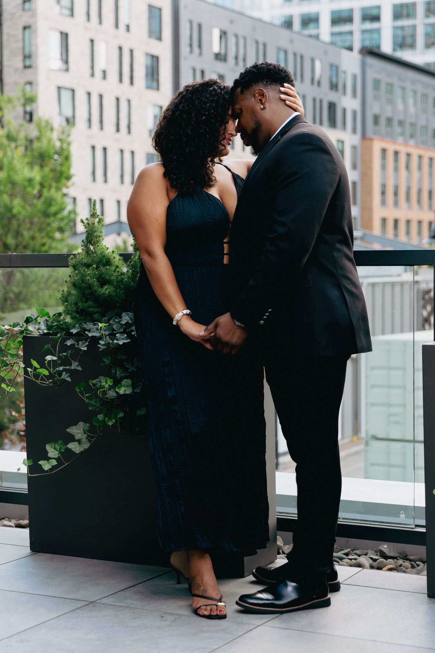 A couple in formal attire shares a romantic moment on an urban rooftop terrace surrounded by city buildings.