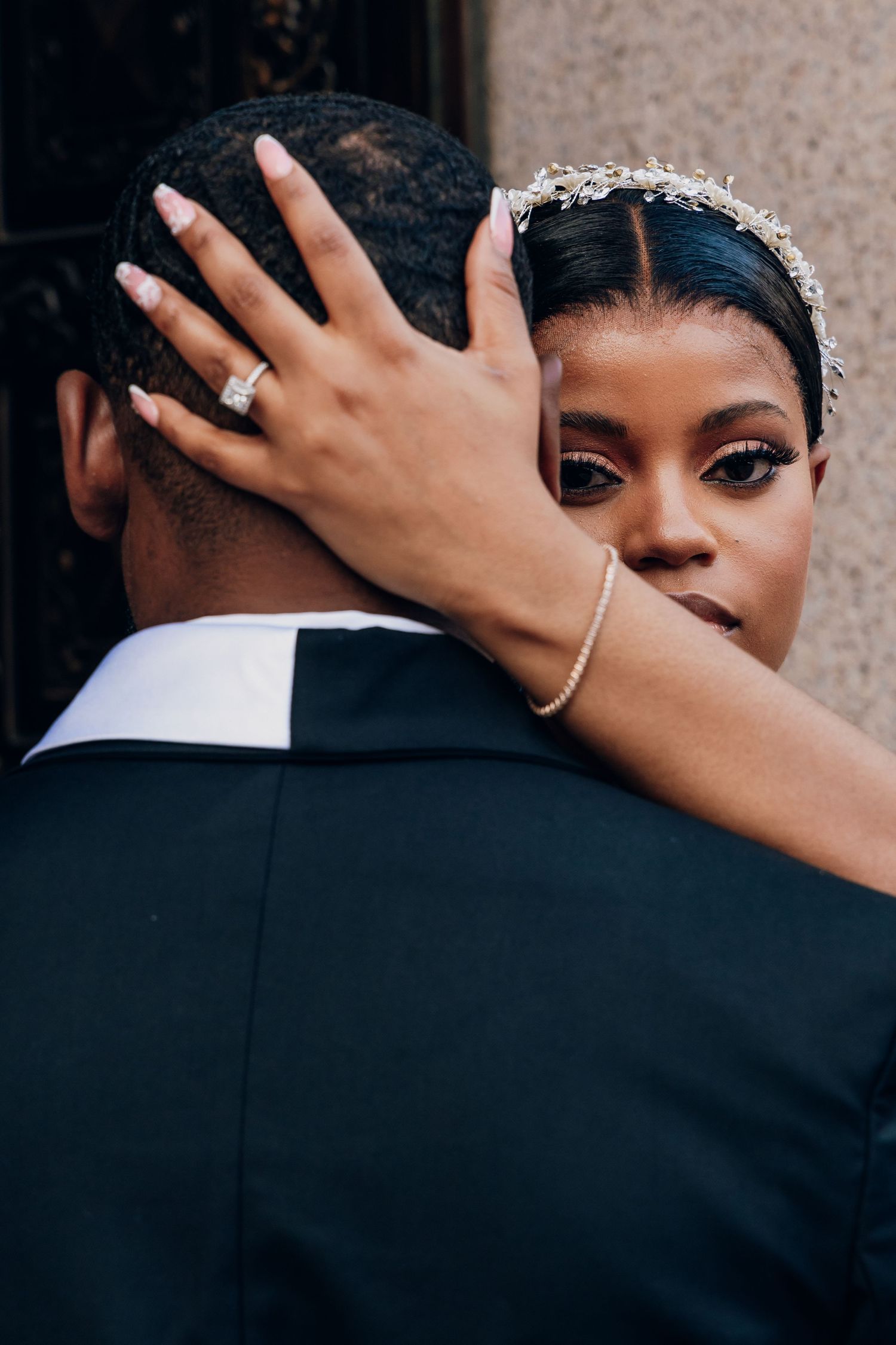 A bride wearing a crystal headband and diamond ring embraces her groom in a black suit during a wedding portrait.