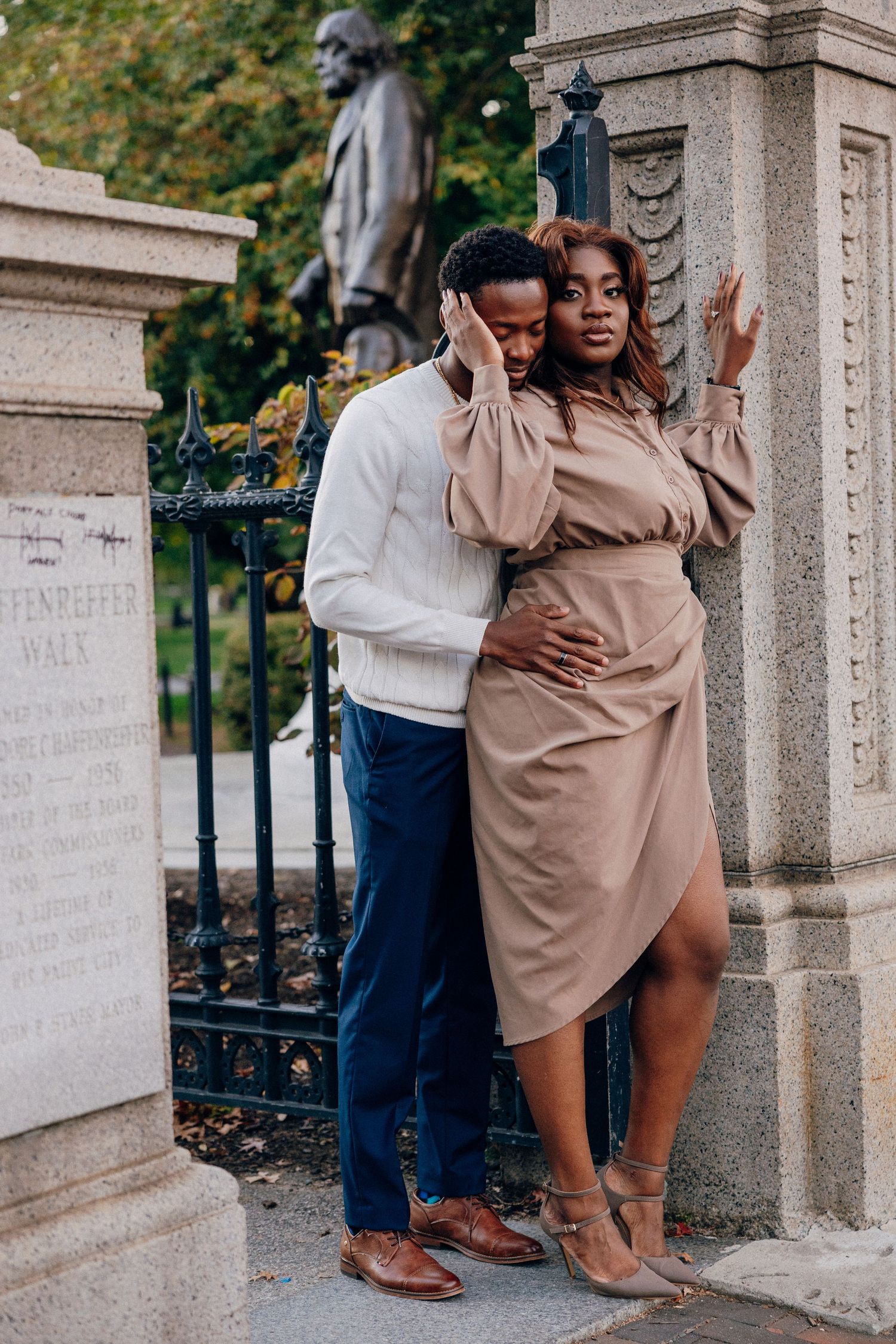 Elegant Black couple posing romantically by a stone monument and iron gate in a lush park setting.