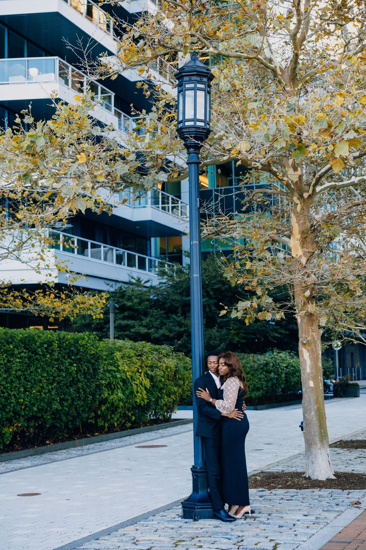 Couple embracing by a vintage street lamp in an urban park with autumn trees and modern building backdrop.
