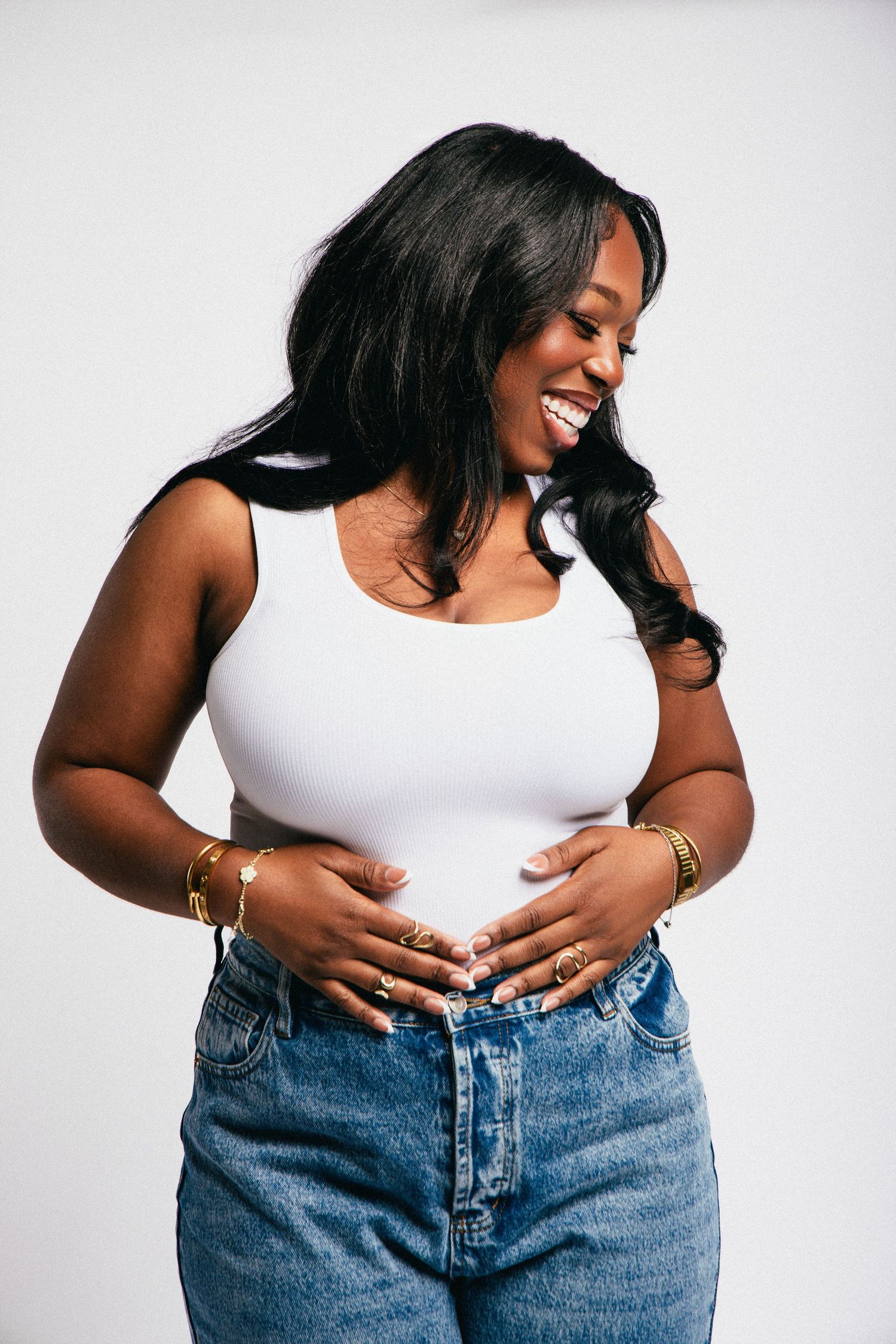 Smiling Black woman in white tank top and jeans, wearing gold jewelry, posing against white background.