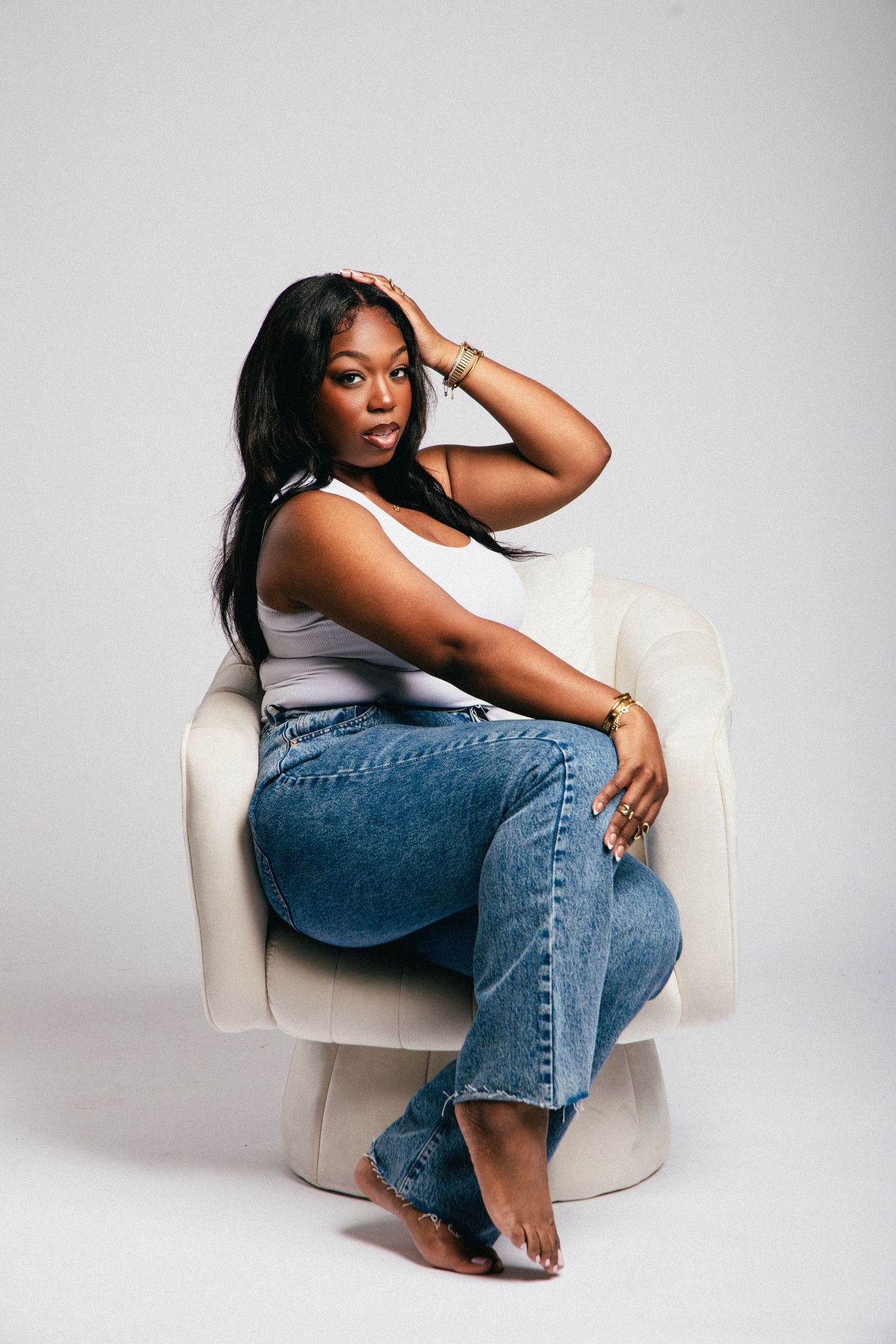 Woman in white tank top and blue jeans sitting in a white chair, posing with hand on head.
