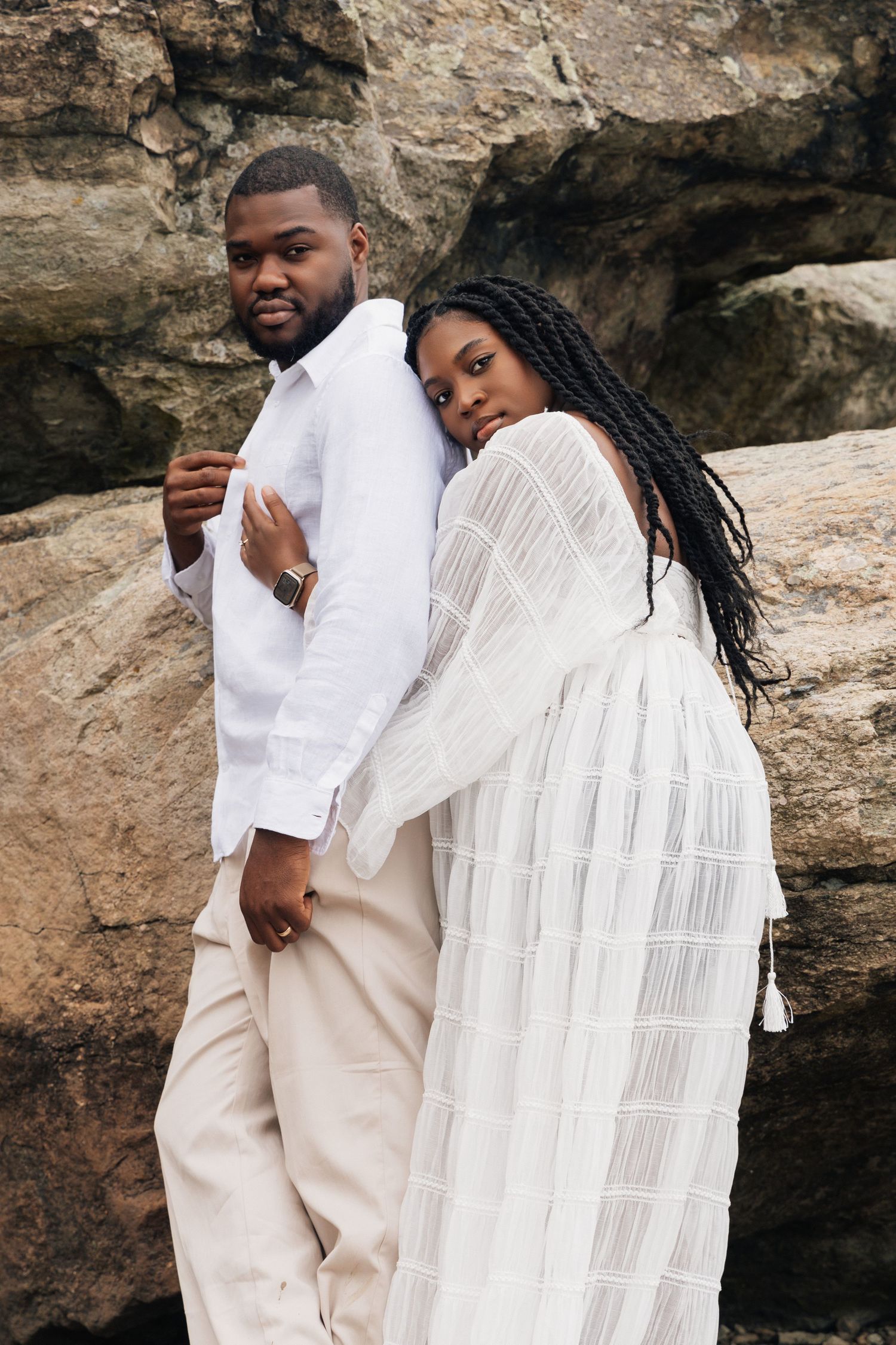 A Black couple in matching white outfits poses together against a rocky backdrop outdoors.