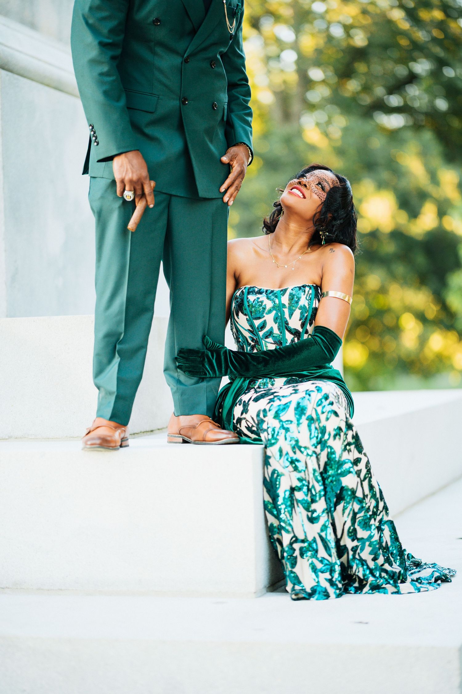 Woman in floral green gown sitting on steps gazing up at man in green suit in a sunny park.