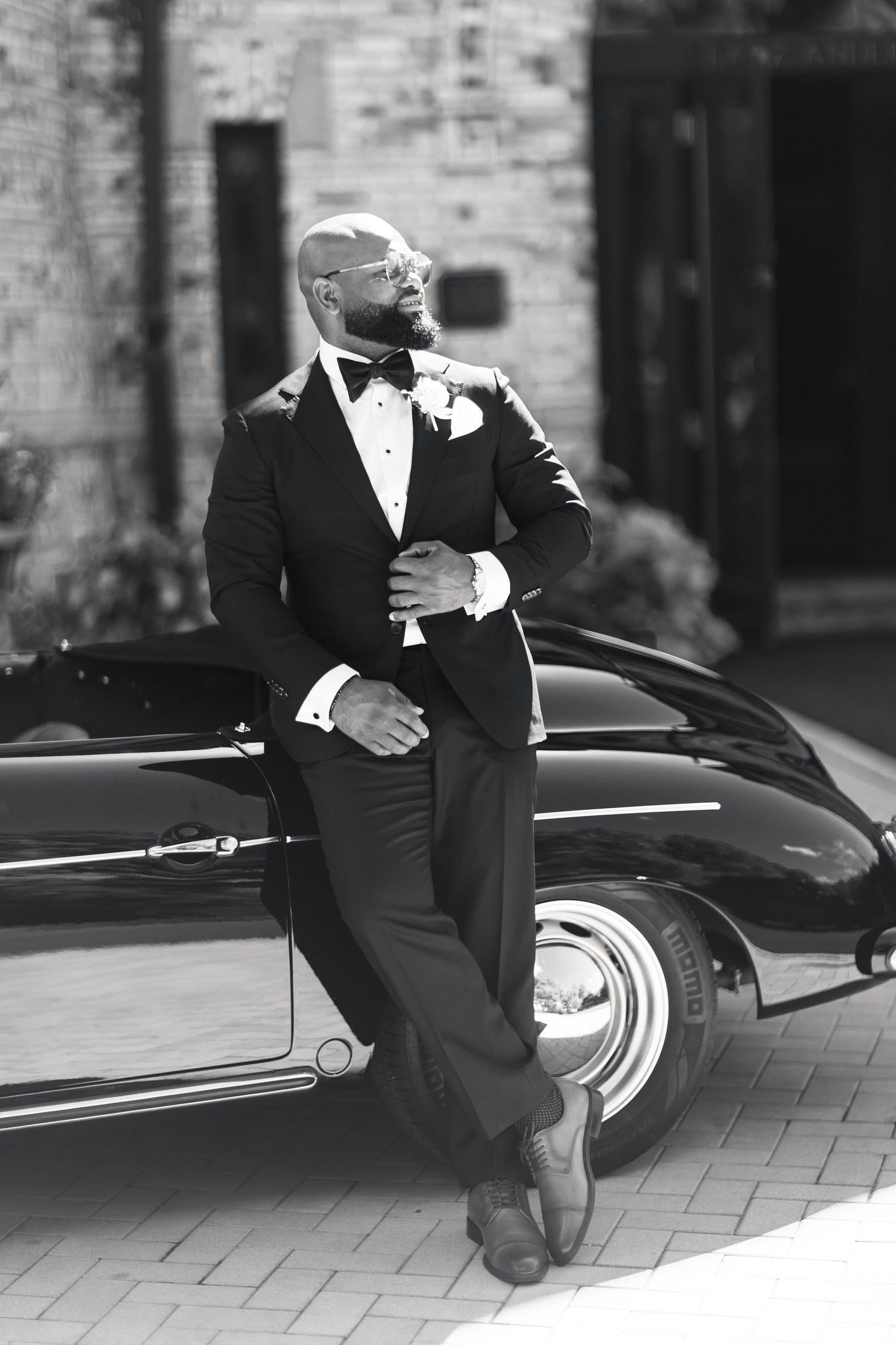 Stylish groom in tuxedo leaning against classic vintage black convertible car in black and white.