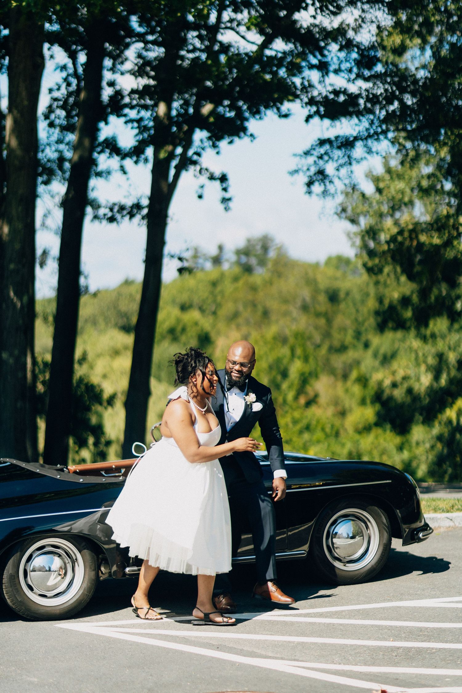Bride and groom share an intimate moment leaning against a vintage black convertible in a sunny tree-lined parking lot.