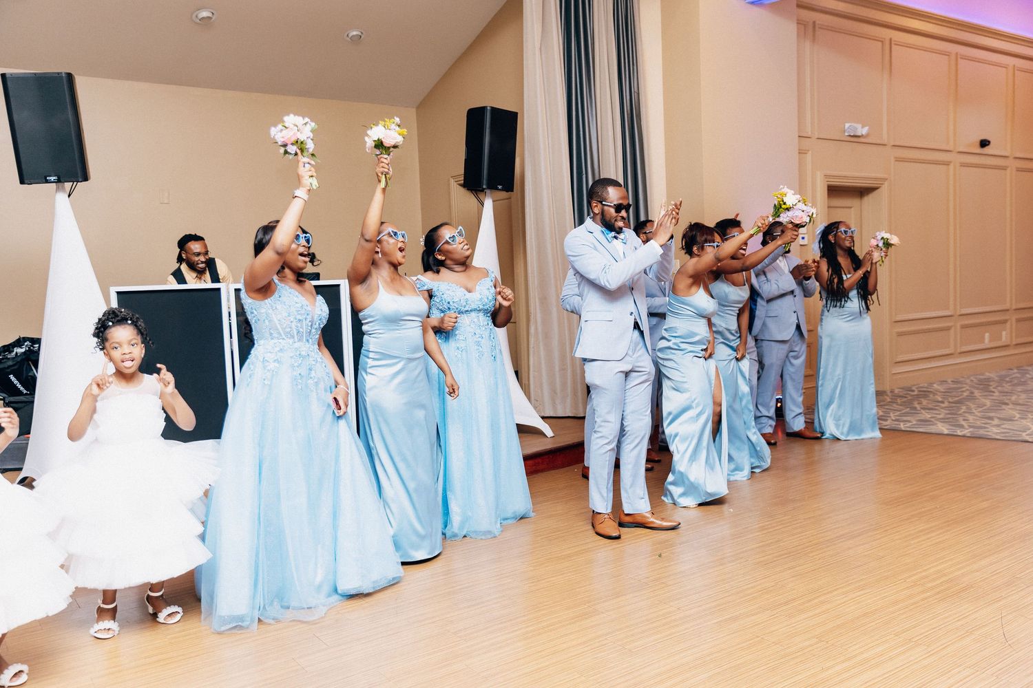 Wedding party in light blue attire dancing joyfully at a reception, bridesmaids holding bouquets aloft.
