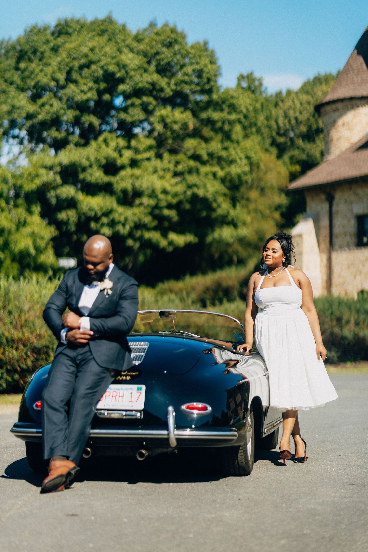 Bride and groom leaning against a classic black Porsche convertible outside a stone building.