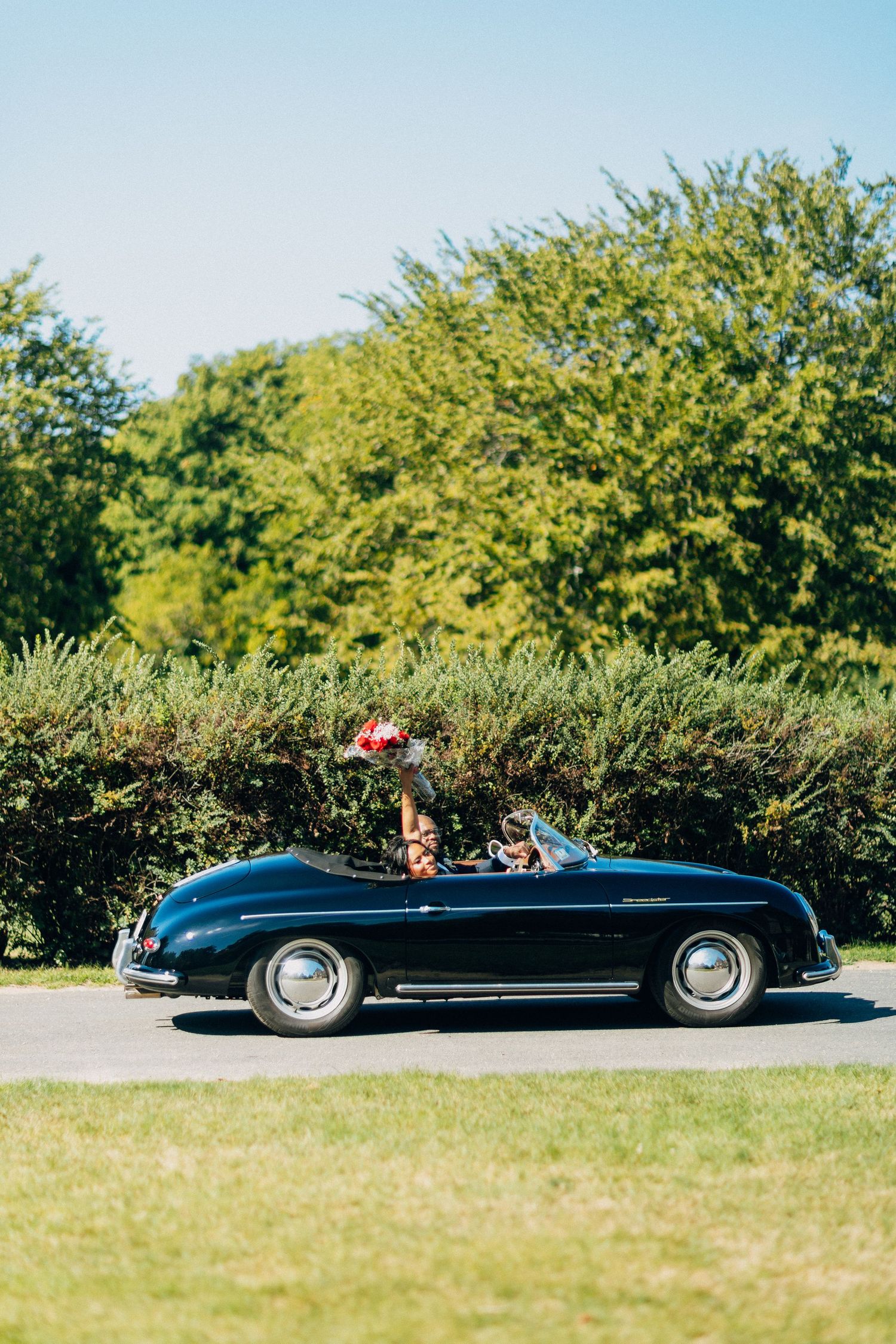 Newlyweds cruising in a vintage black Porsche 356 convertible, bride raising bouquet joyfully.