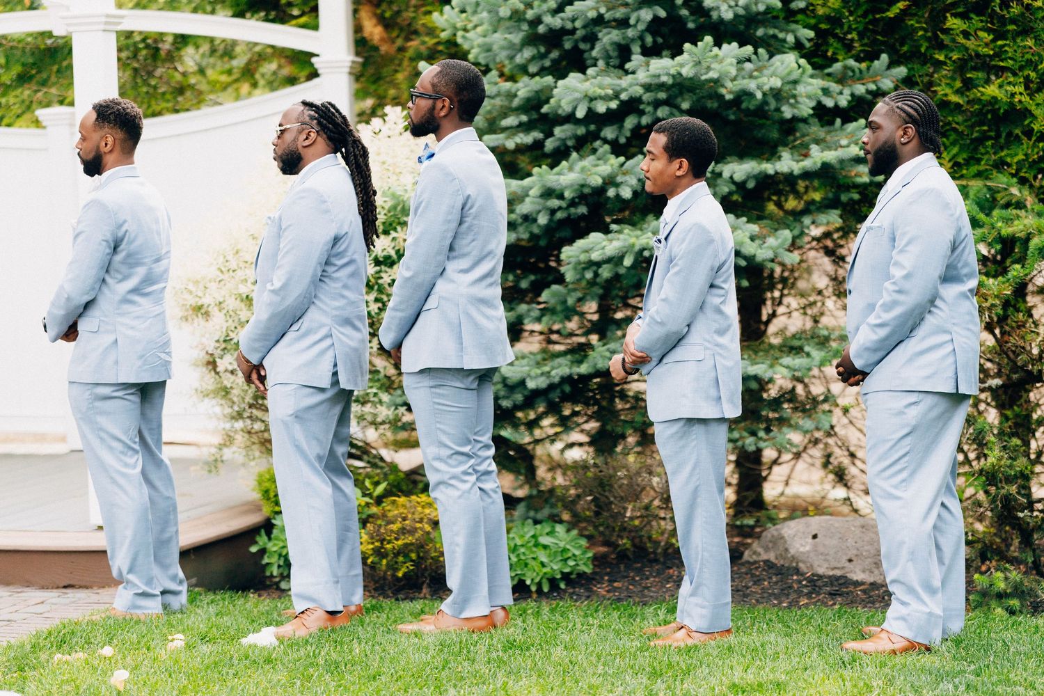 Four groomsmen in matching light blue suits stand in line outdoors at wedding ceremony.