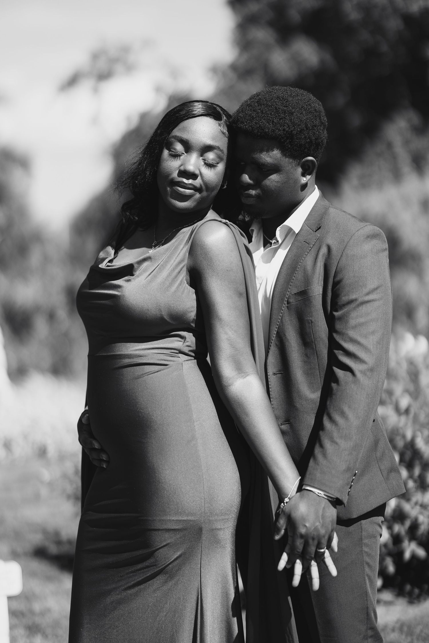Black and white photo of a couple embracing outdoors, woman in elegant dress, man in suit holding hands tenderly.