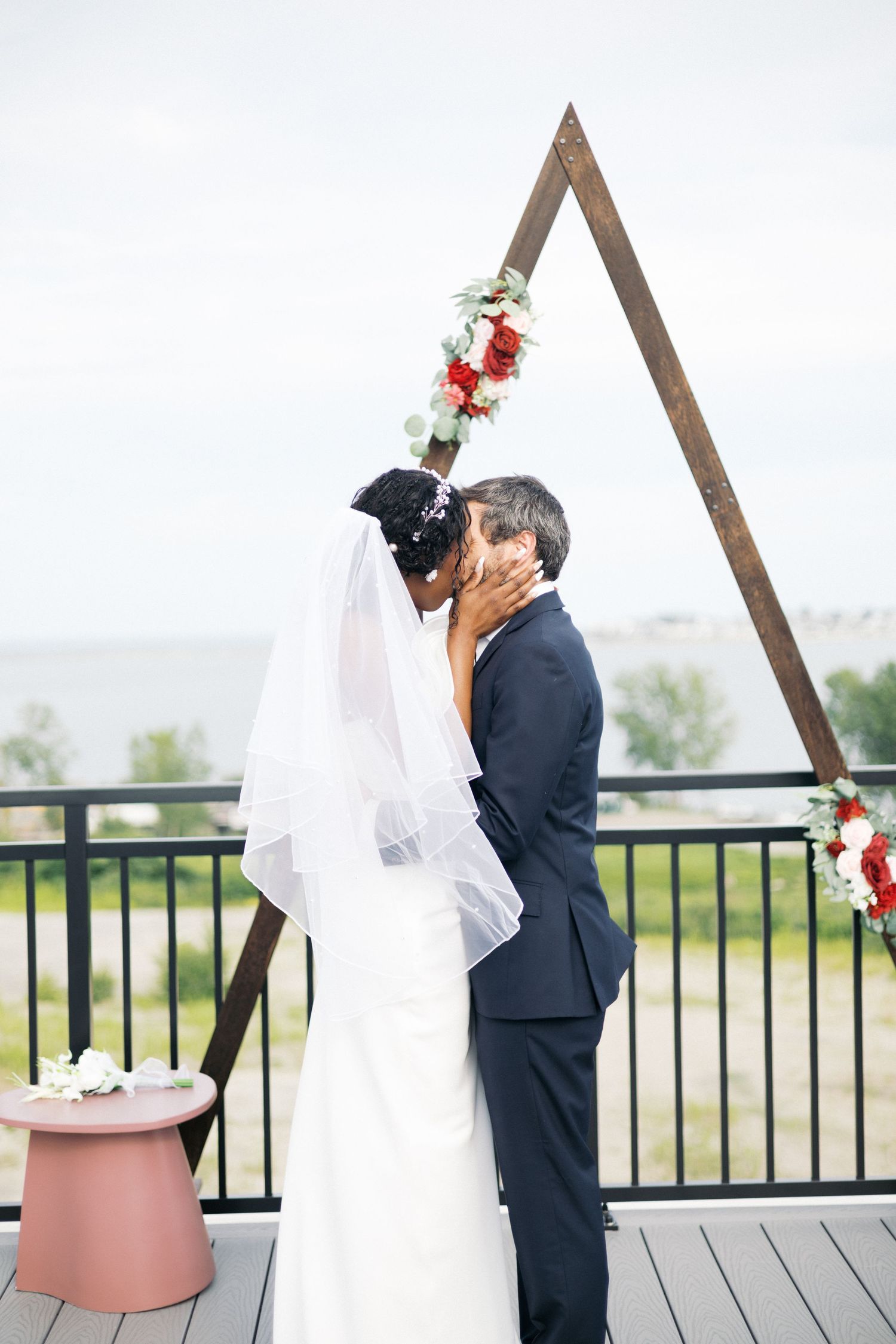 An interracial bride and groom share their first kiss under a triangular wooden arch with floral decor.