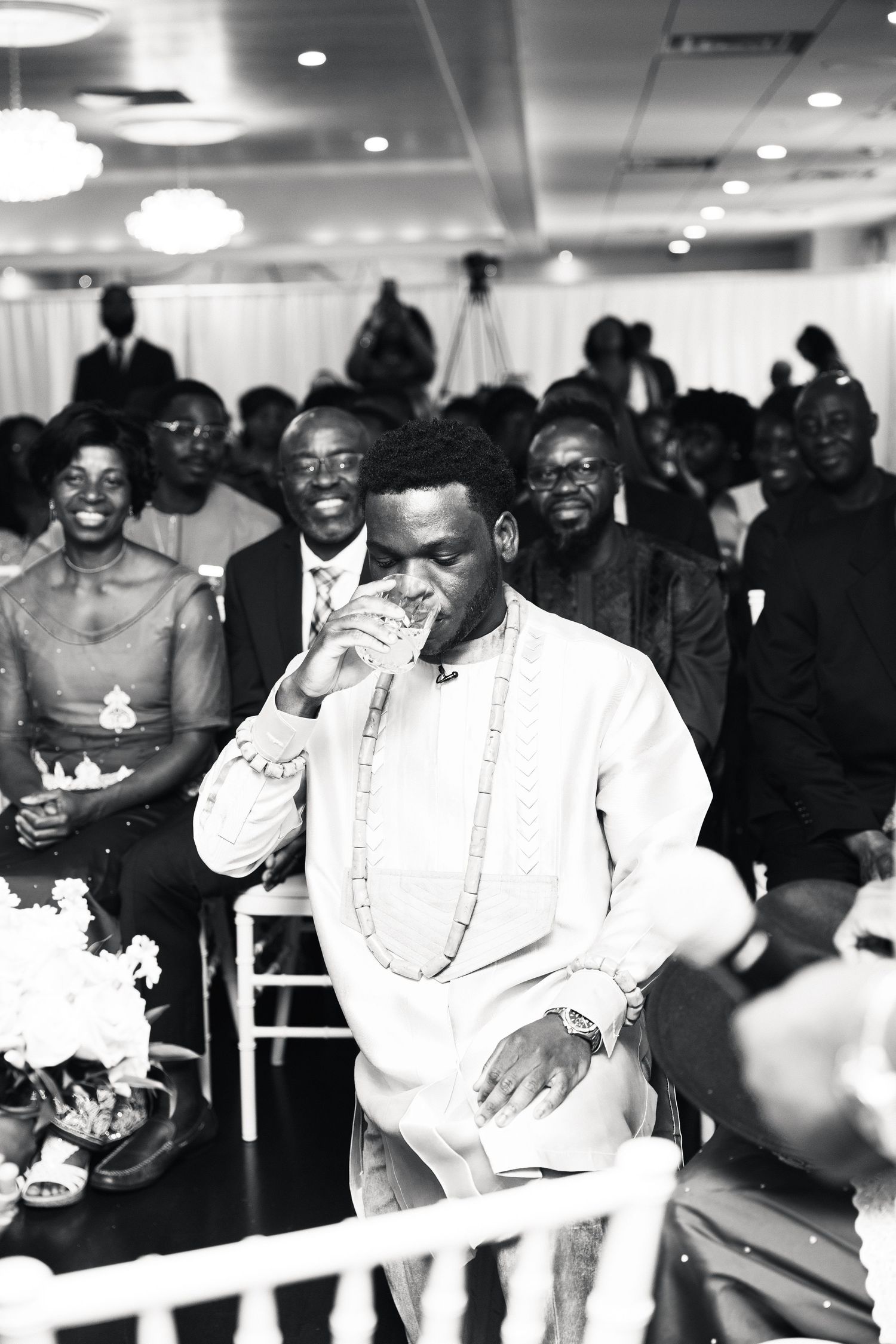 Groom in white traditional African attire drinks from glass during wedding ceremony while guests smile in background.
