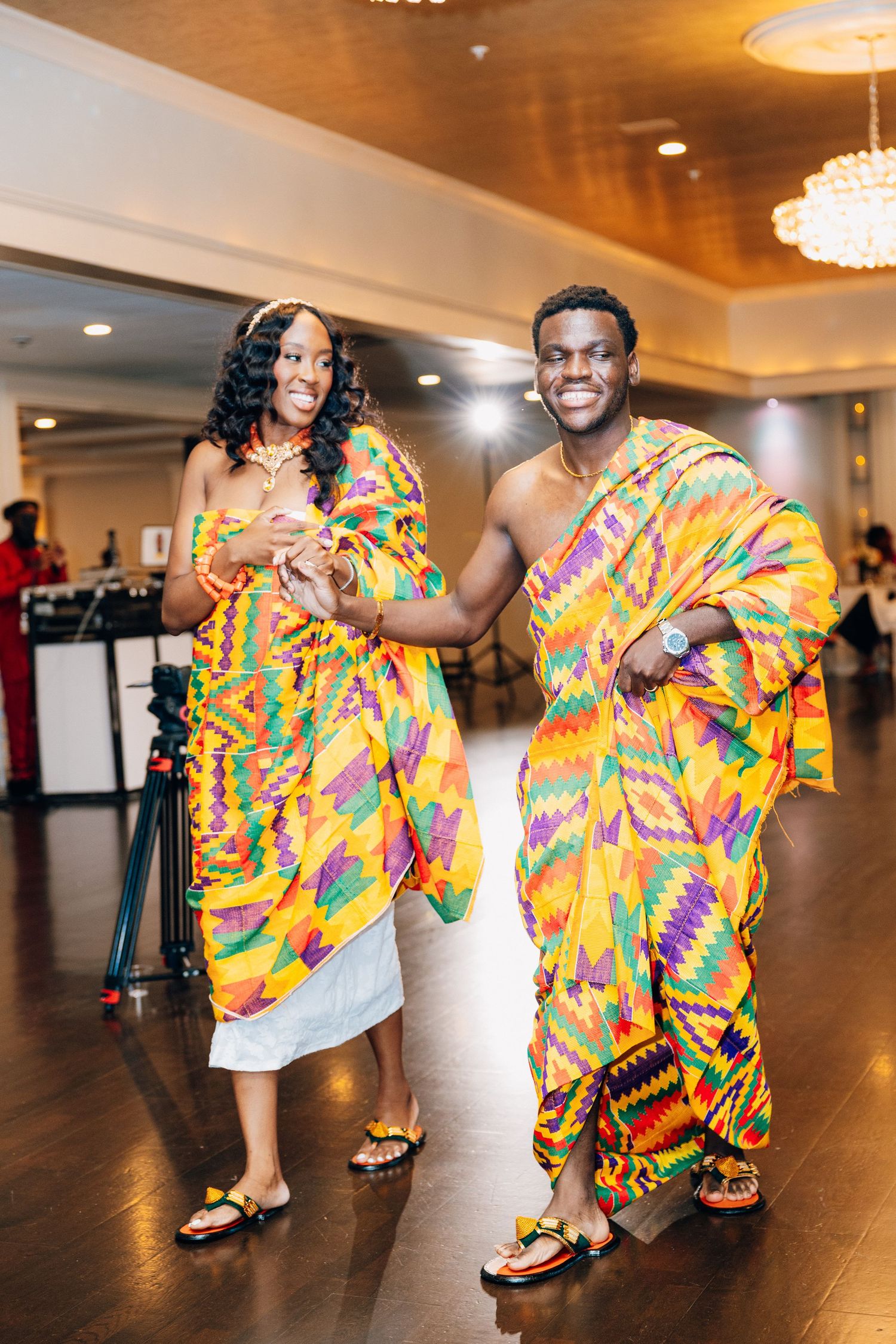 Smiling couple posing together in vibrant kente cloth traditional African attire at a wedding celebration.