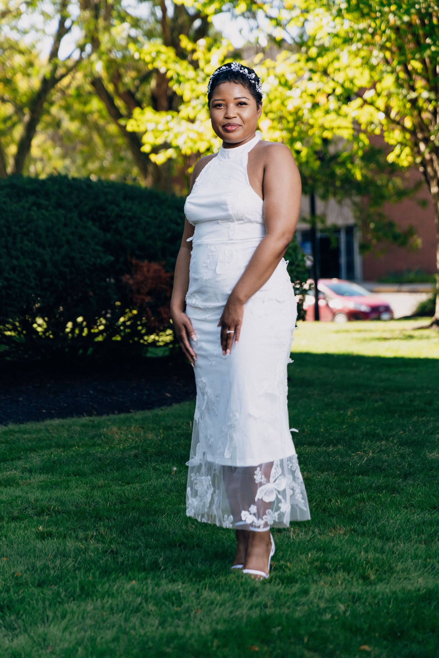 A bride in a white lace midi dress and floral headpiece poses on a green lawn outdoors.