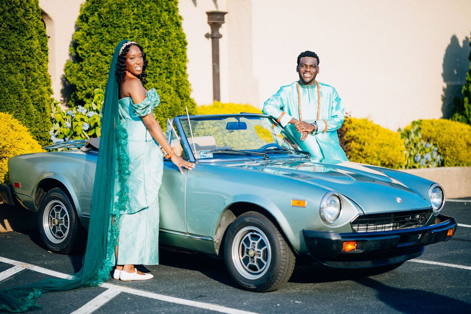 Bride and groom in matching teal outfits posing beside a vintage silver convertible car.