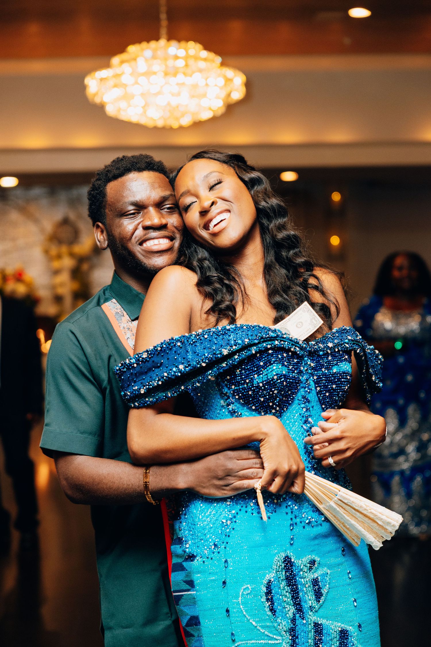 A smiling couple in African traditional attire embraces warmly under a chandelier at a celebratory event.