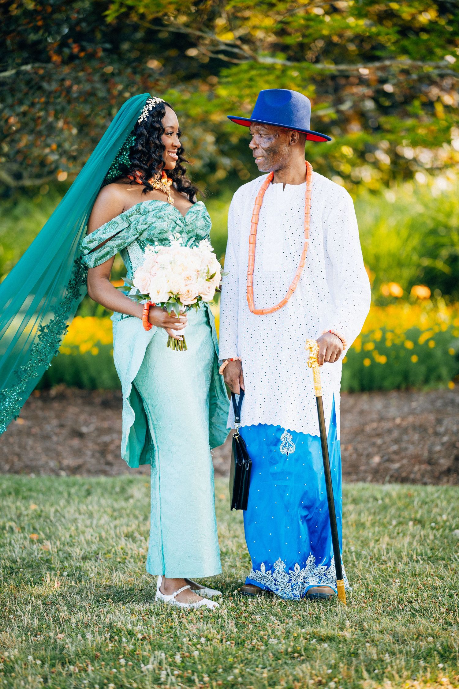 Nigerian bride in mint green gown with teal veil and groom in white agbada with blue hat at outdoor wedding.