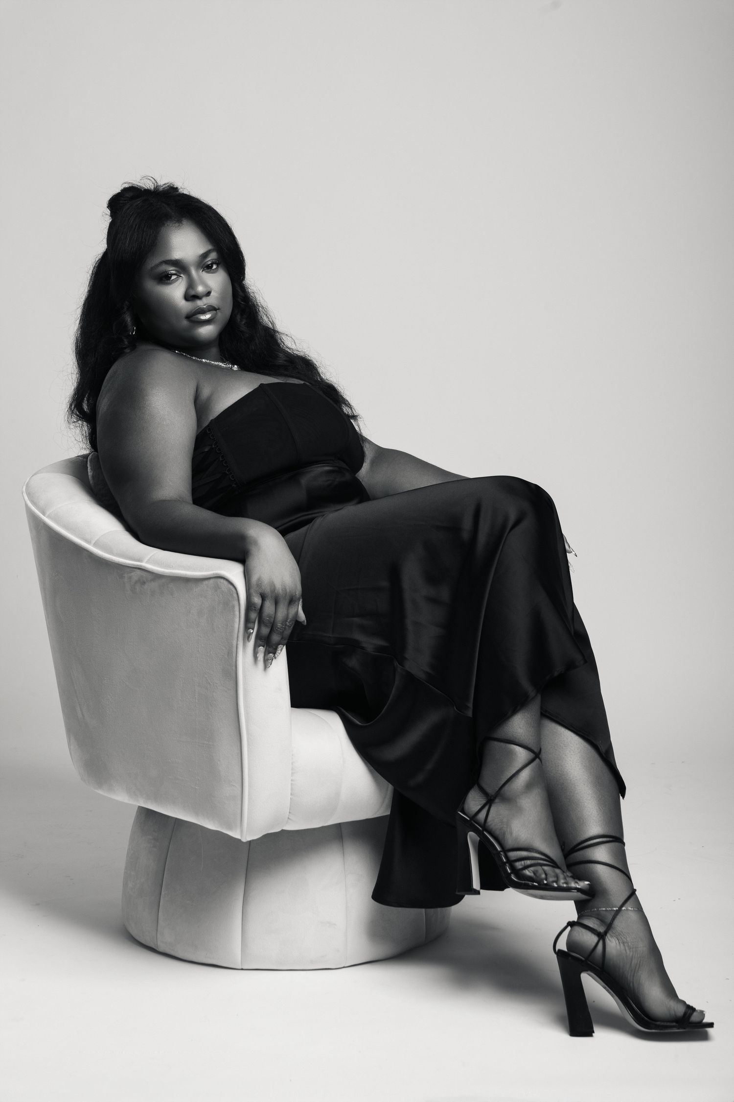 Black and white portrait of a woman in a black strapless outfit seated in a white chair with heels.