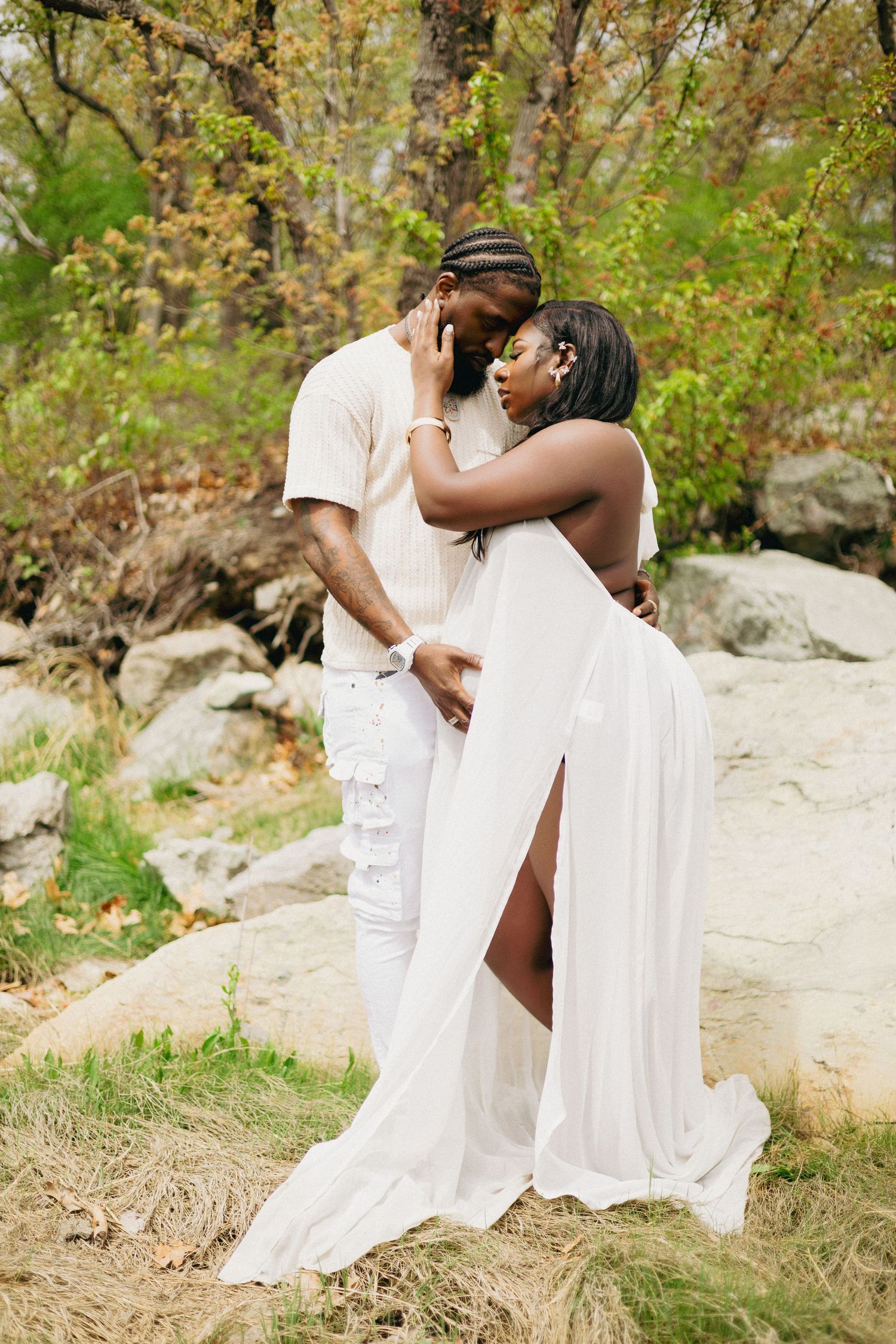 A couple in white outfits share a romantic moment outdoors among rocks and autumn trees.