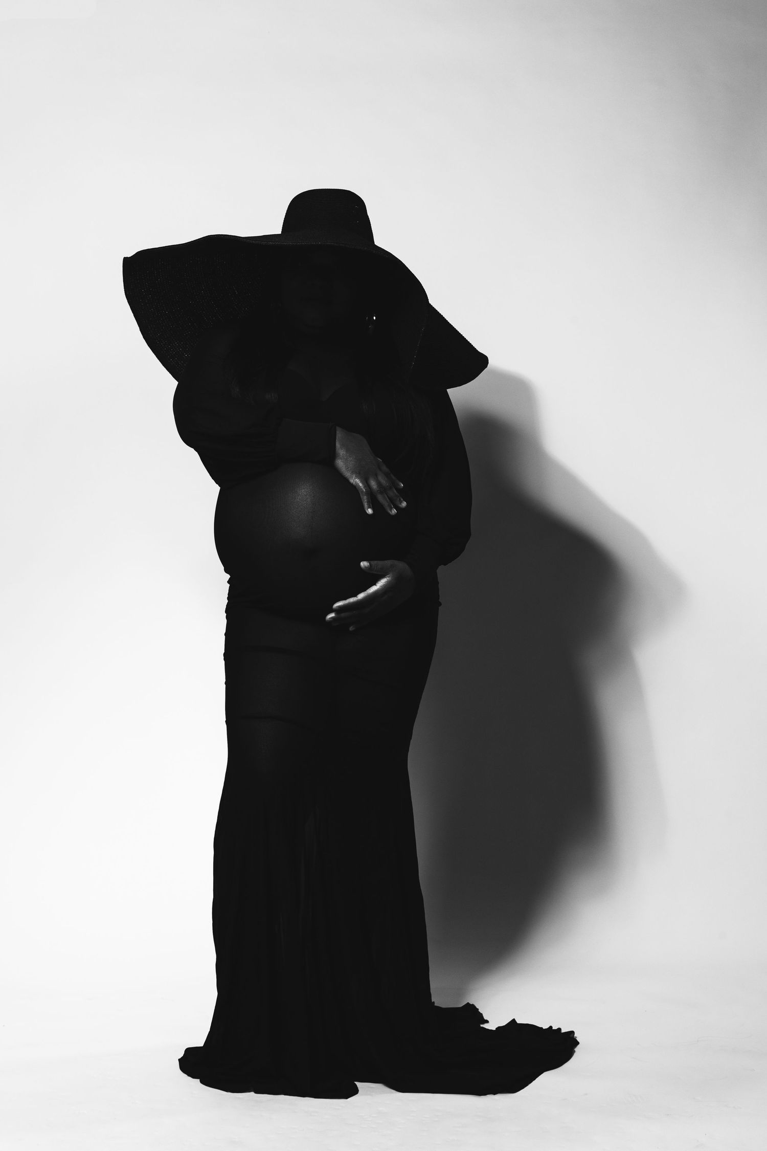 Black and white maternity photo of pregnant woman in elegant black gown and wide-brim hat cradling her baby bump.
