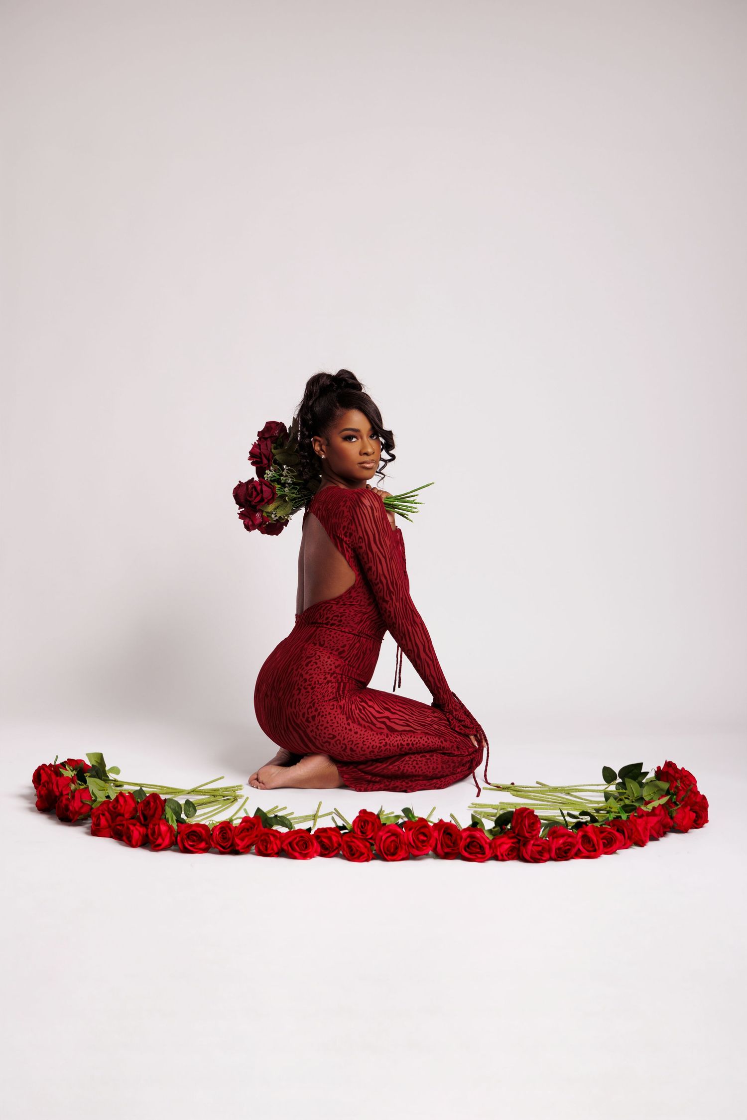Woman in red sequin gown sits surrounded by red roses in a elegant studio photo shoot.