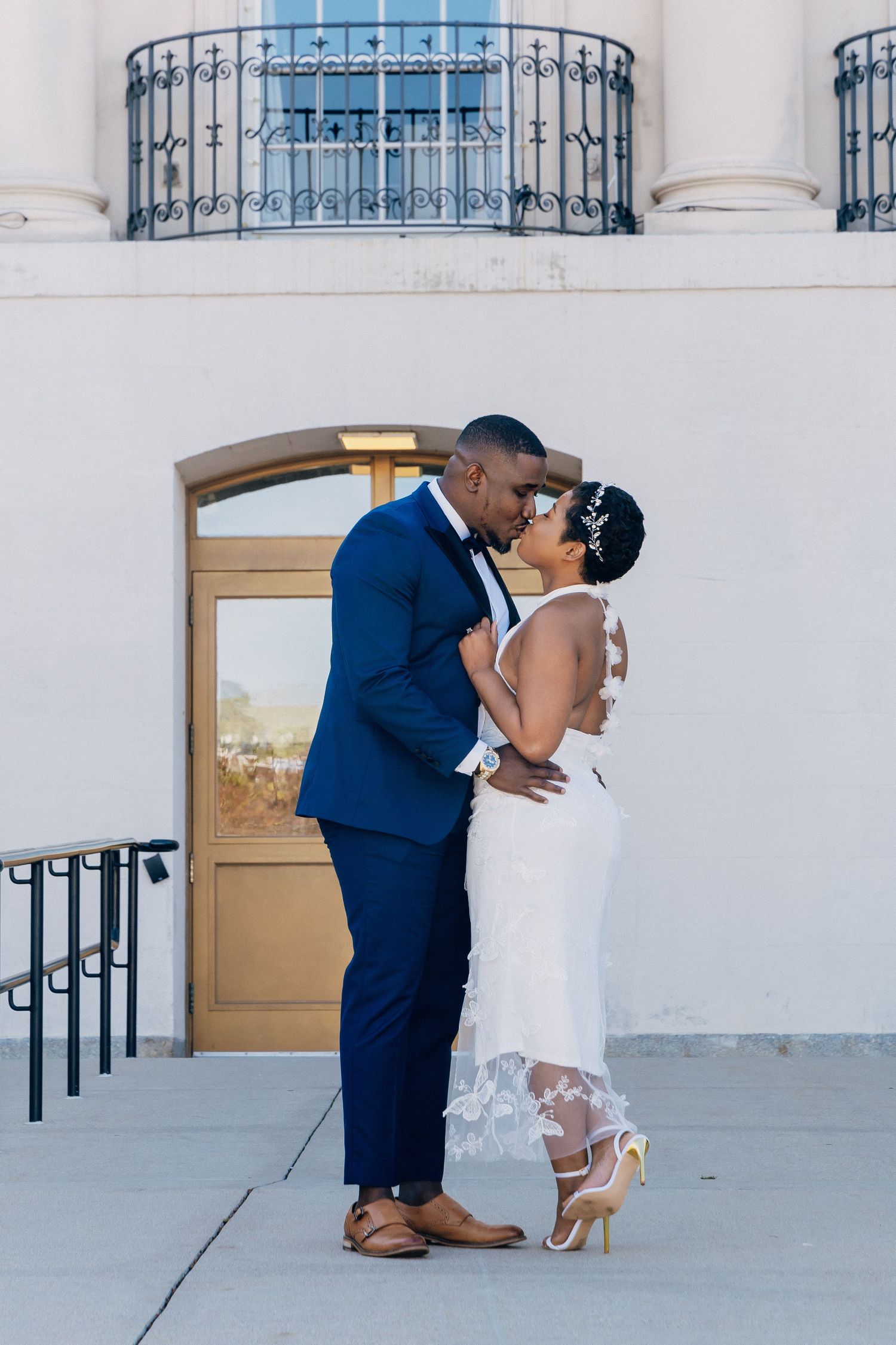 Bride and groom share a romantic kiss outside historic building, groom in navy tuxedo, bride in lace gown.