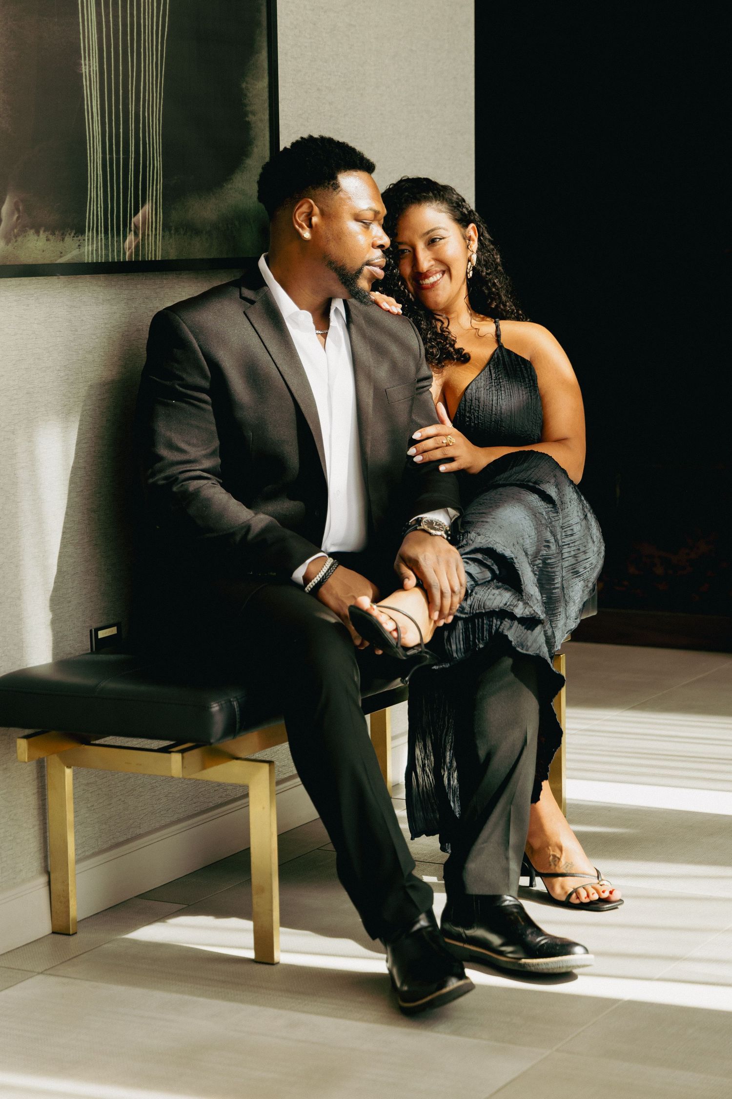 Elegant couple sitting together in sunlight, man in suit and woman in black dress, sharing an intimate moment.