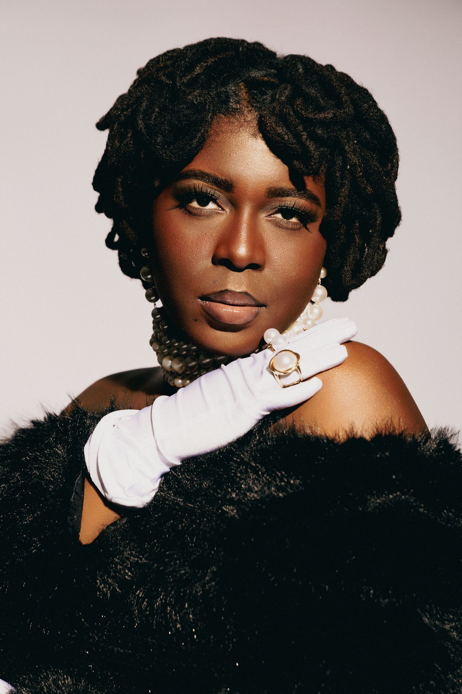Elegant Black woman in fur coat, white gloves, gold ring, and locs hairstyle poses confidently against gray background.