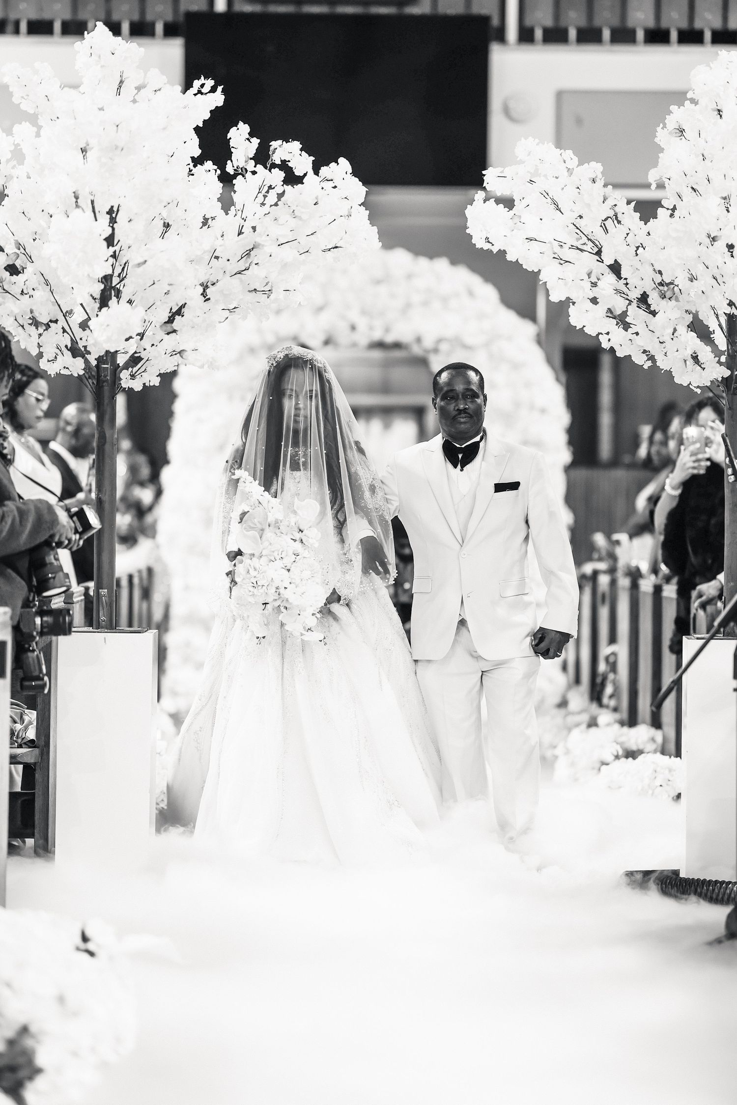 Black and white photo of bride and groom walking down the aisle at elegant church wedding with white floral arch.