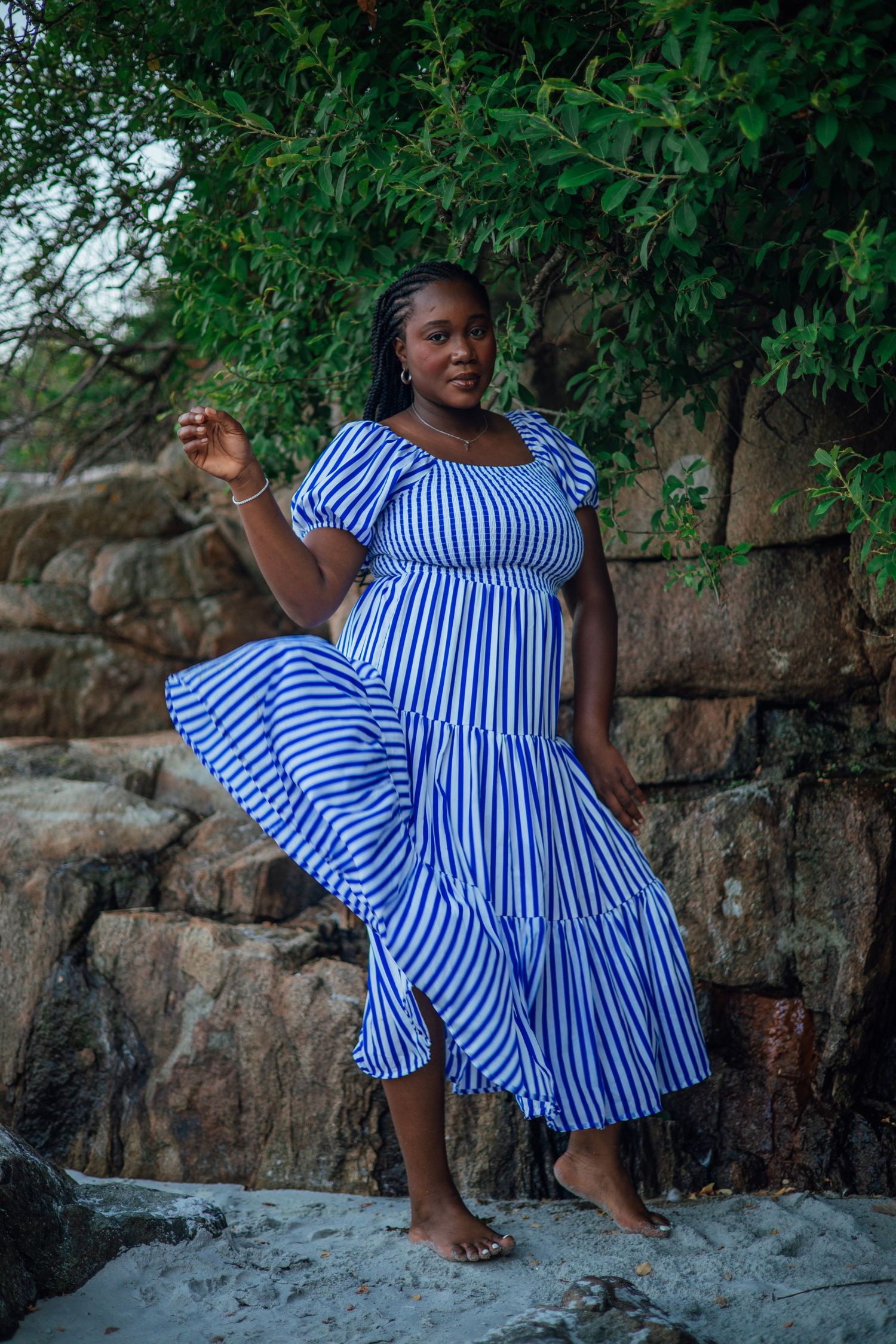 A confident woman in a blue and white striped maxi dress stands barefoot on sandy rocks near green trees.