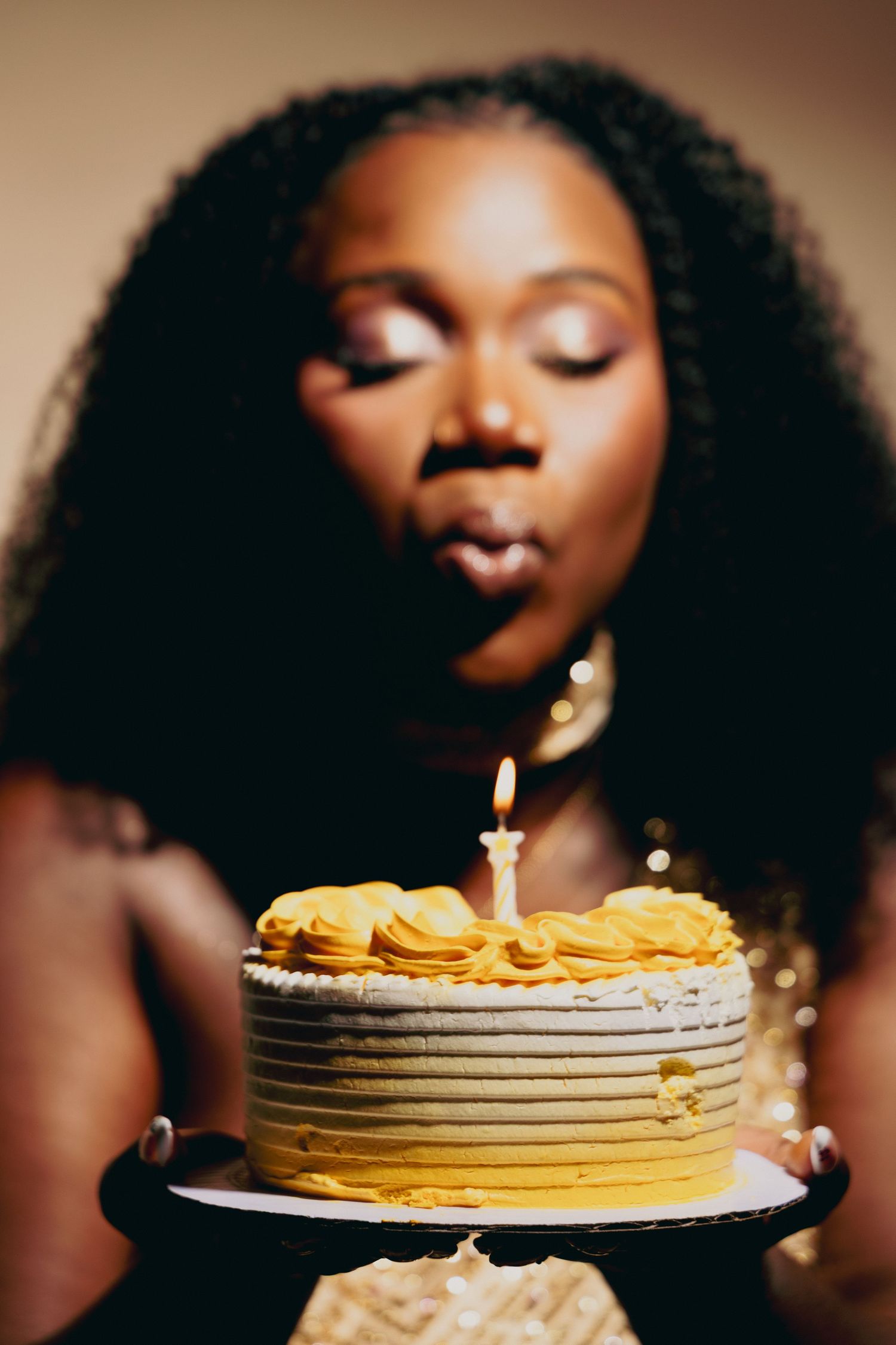 A Black woman with curly hair blows out a candle on a yellow birthday cake she holds on a cake stand.