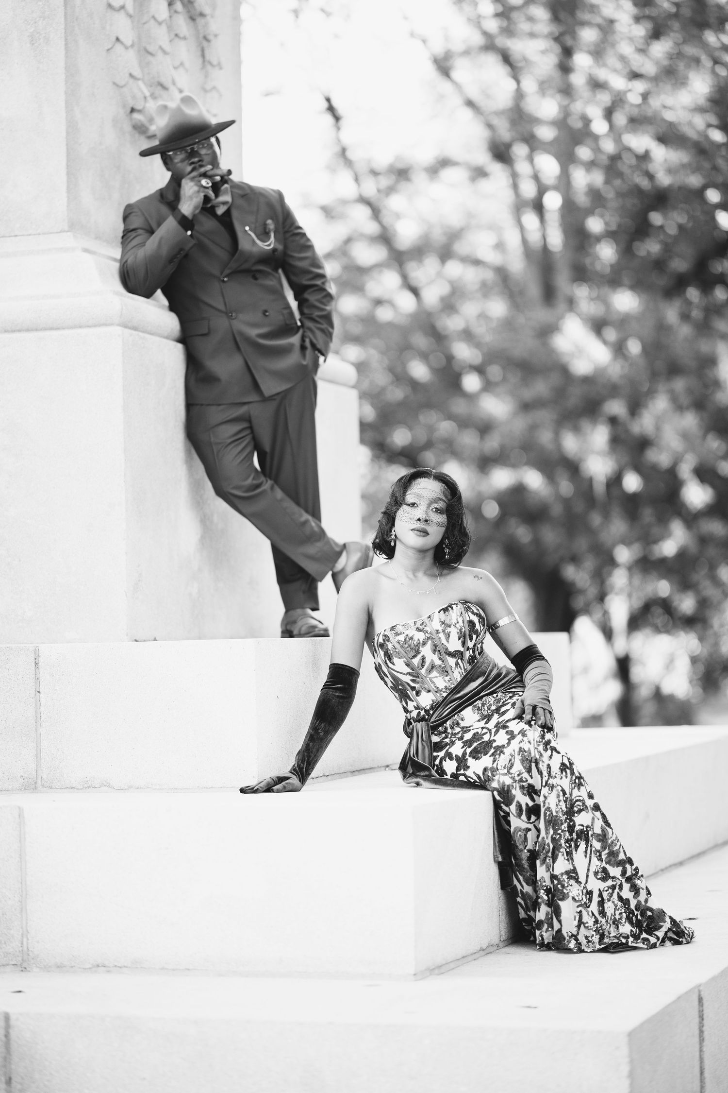 Couple posing on white marble steps, woman in floral gown, man in suit and hat.