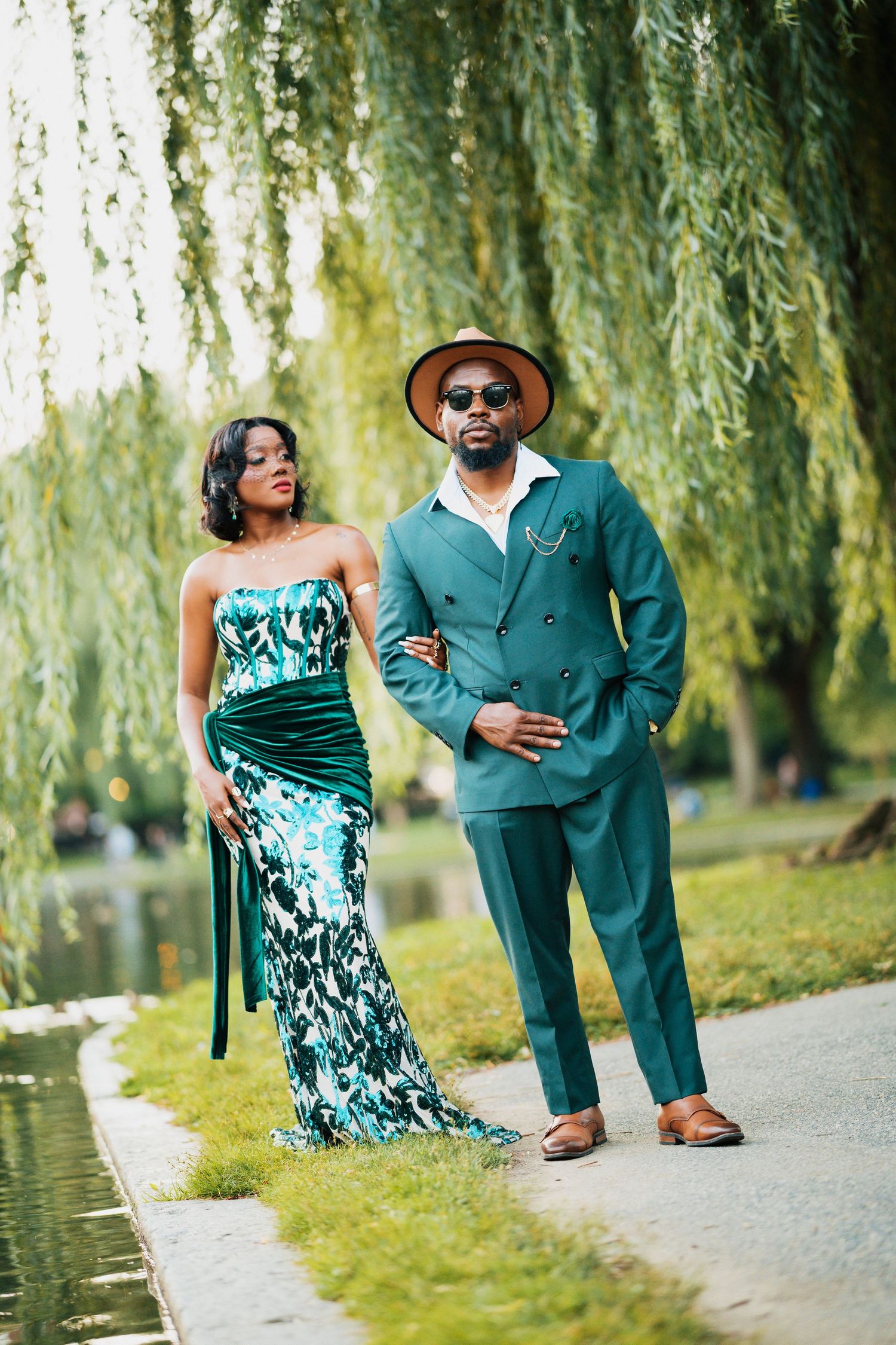 Stylish Black couple in matching emerald green outfits pose near a willow tree by a park pond.