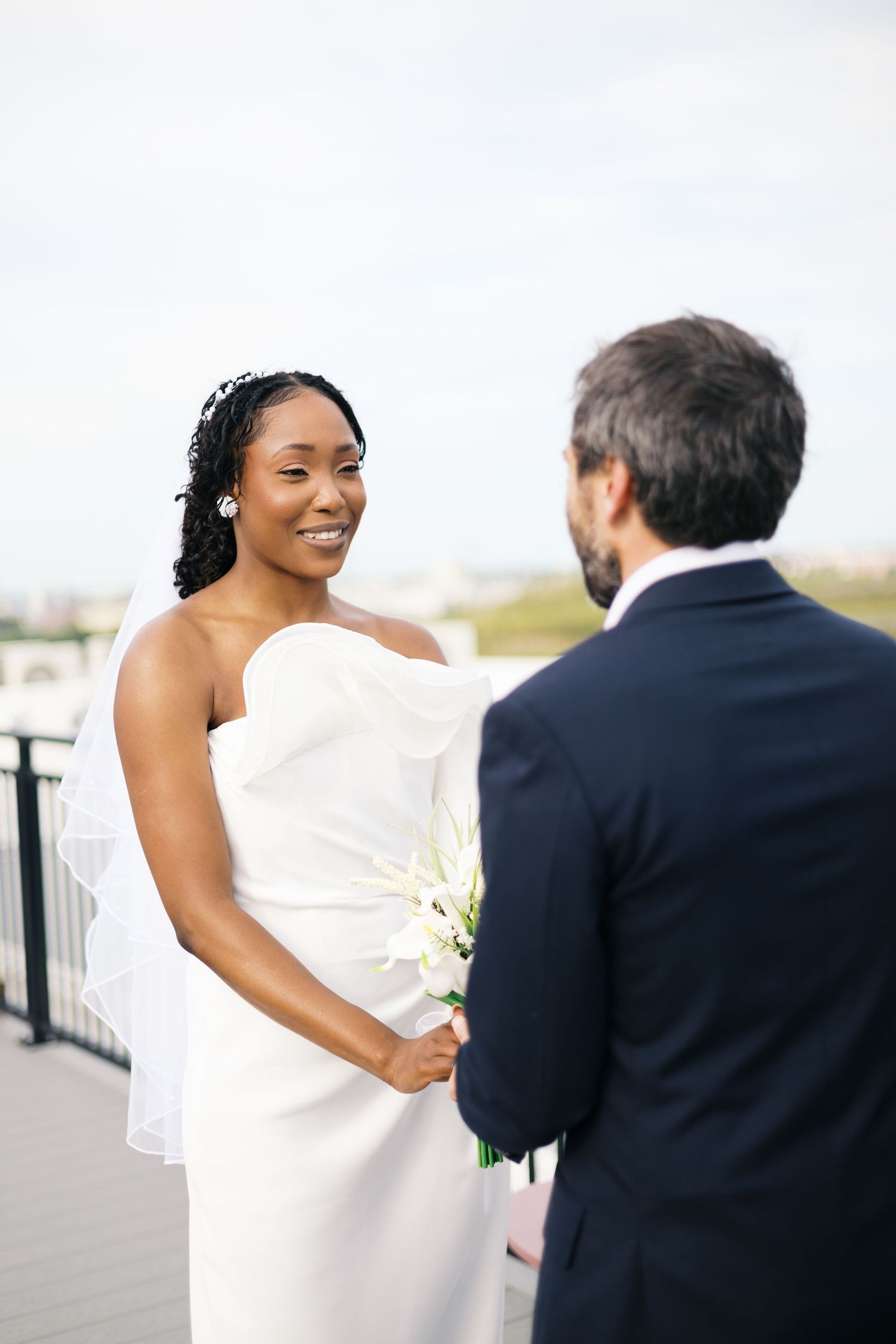 Smiling Black bride in white strapless gown holds white calla lilies while exchanging vows with groom in navy suit outdoors.