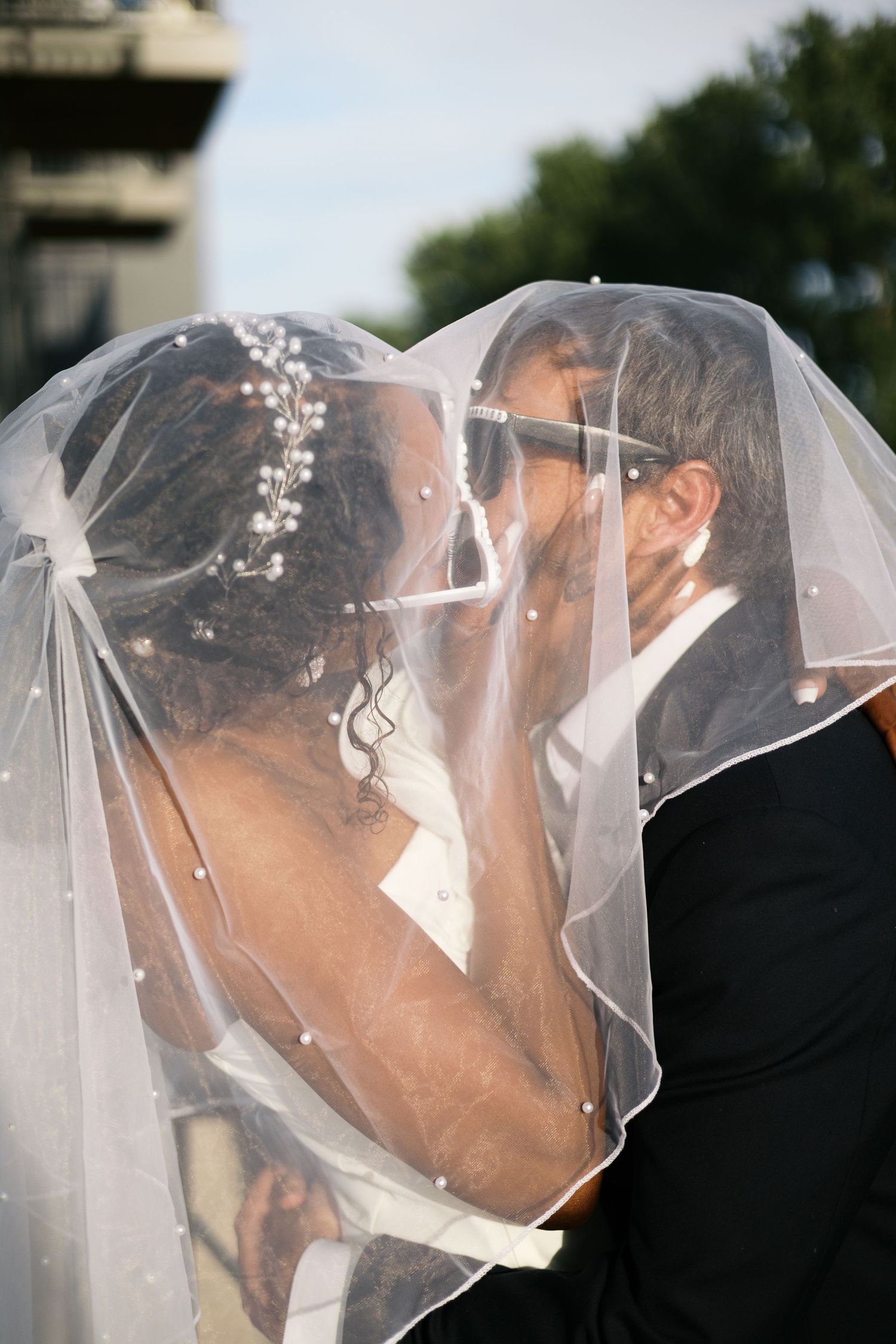 Interracial bride and groom share a romantic kiss beneath a pearl-adorned wedding veil outdoors after their ceremony.